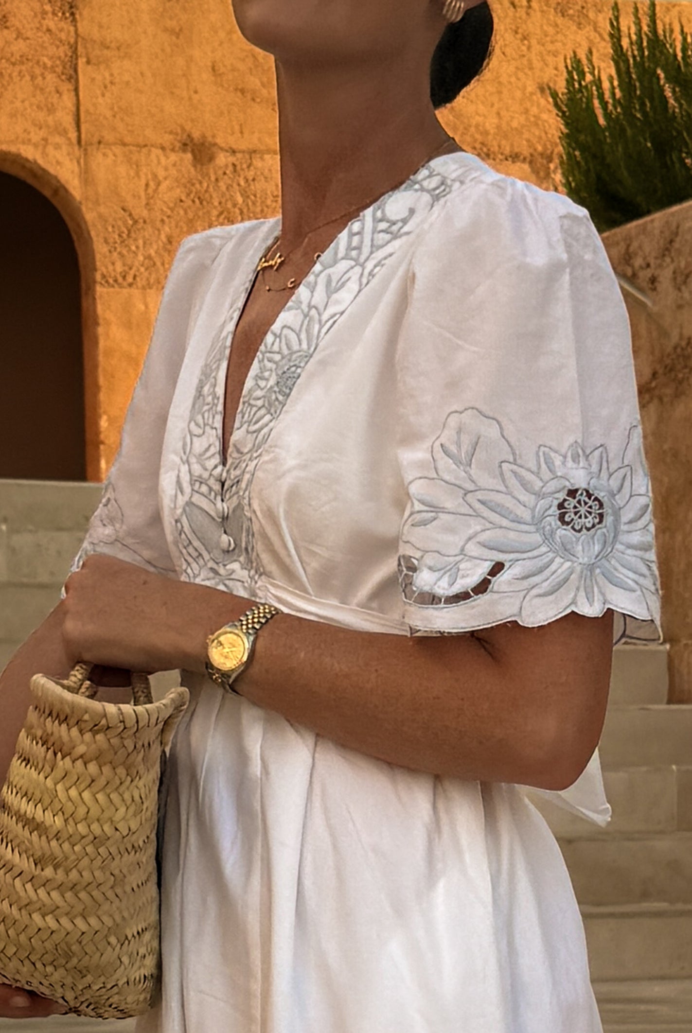 A woman wearing a white embroidered dress with blue floral embroidery on the bodice and sleeve, holding a woven straw bag in front of a warm beige stone wall and steps.