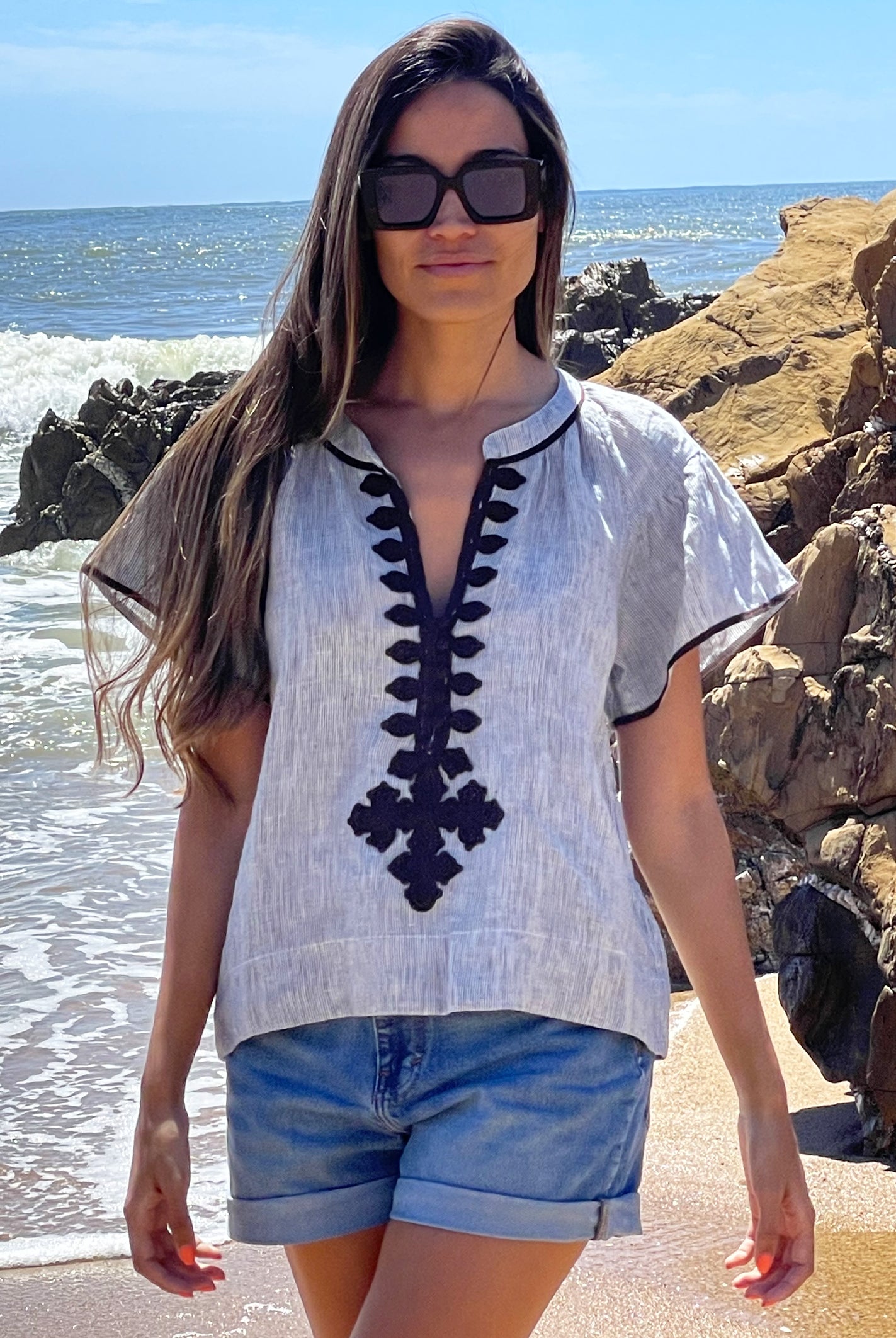 A model on a beach wearing a Rose and Rose striped linen Imperia top, denim shorts and sunglasses. 