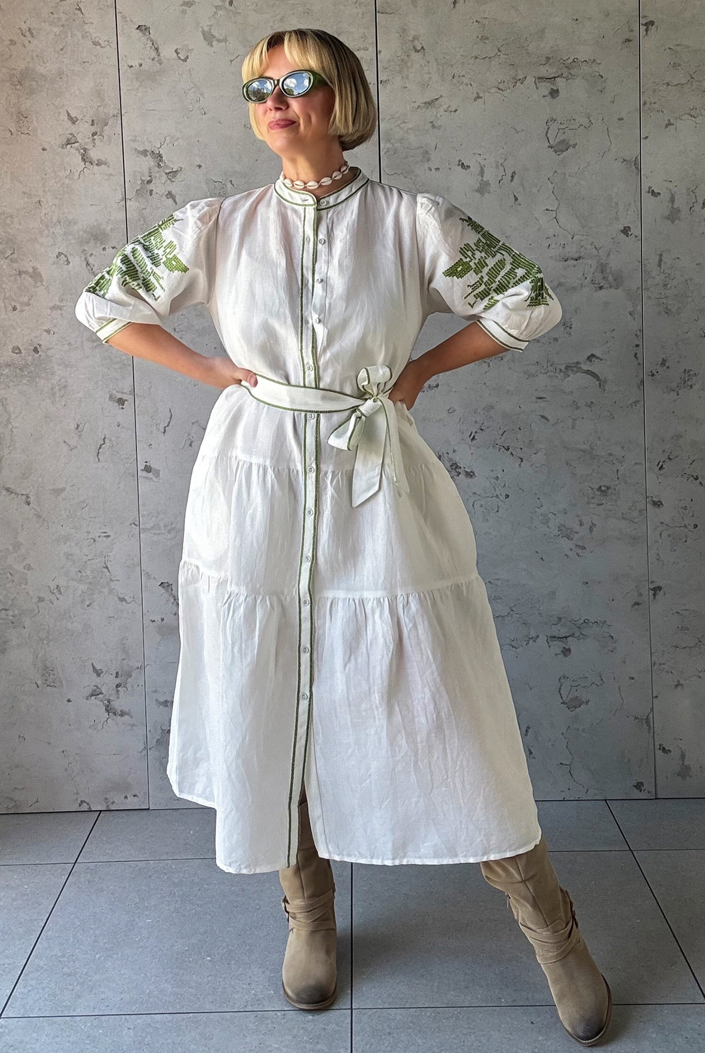 Woman wearing a white dress with green embroidery against a gray tiled wall.
