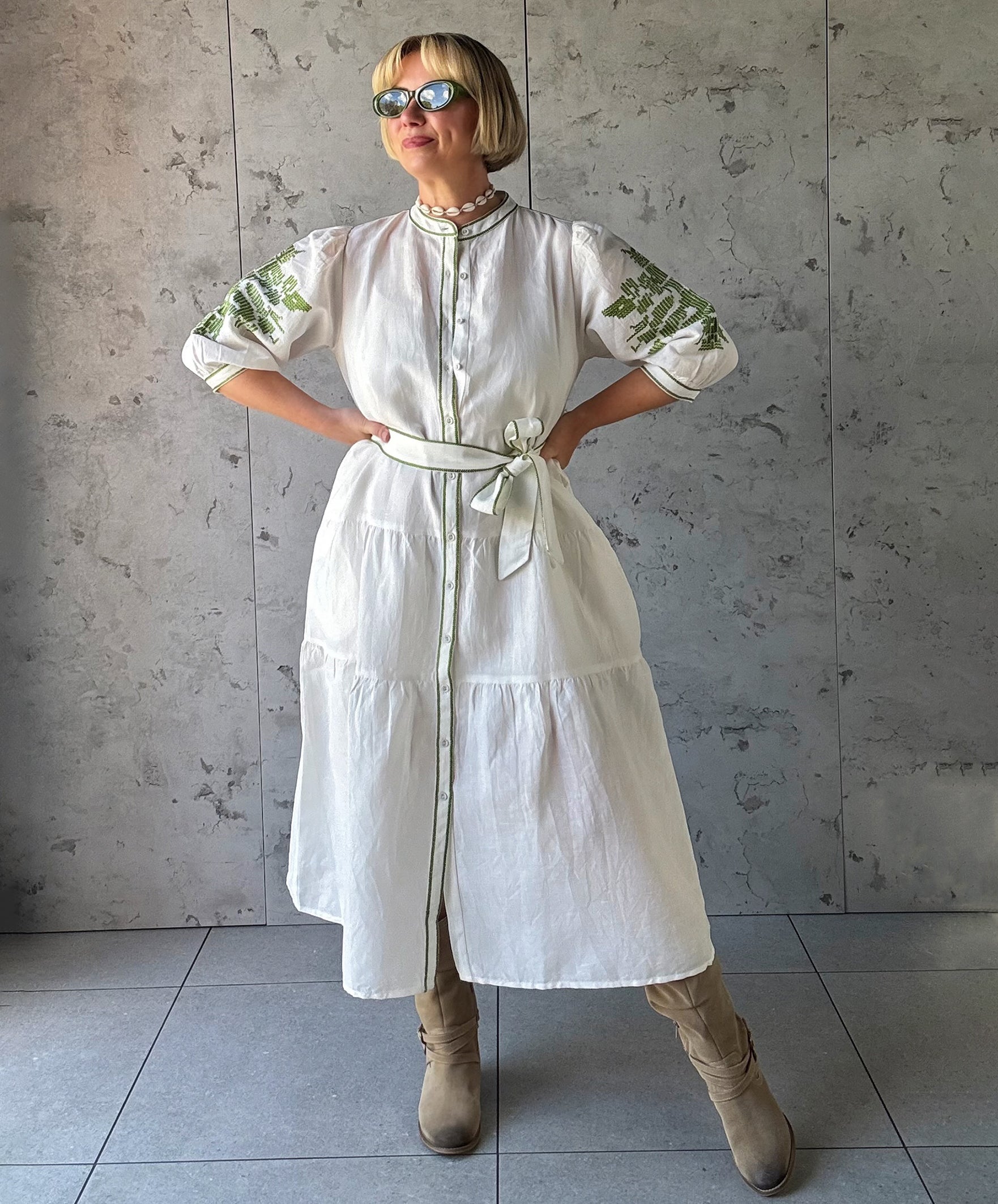 Woman wearing a white dress with green embroidery against a gray tiled wall.