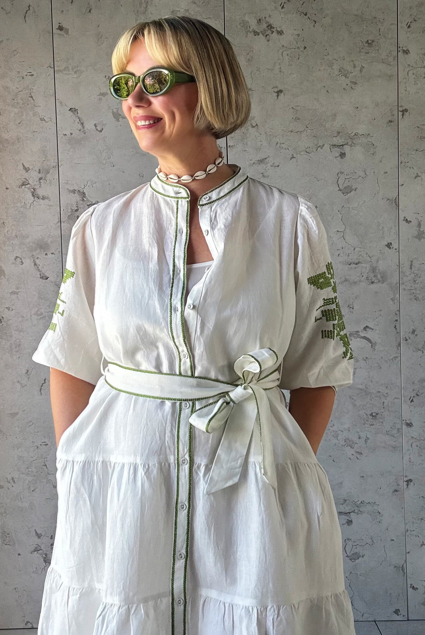 Woman wearing a white dress with green embroidery against a gray wall.