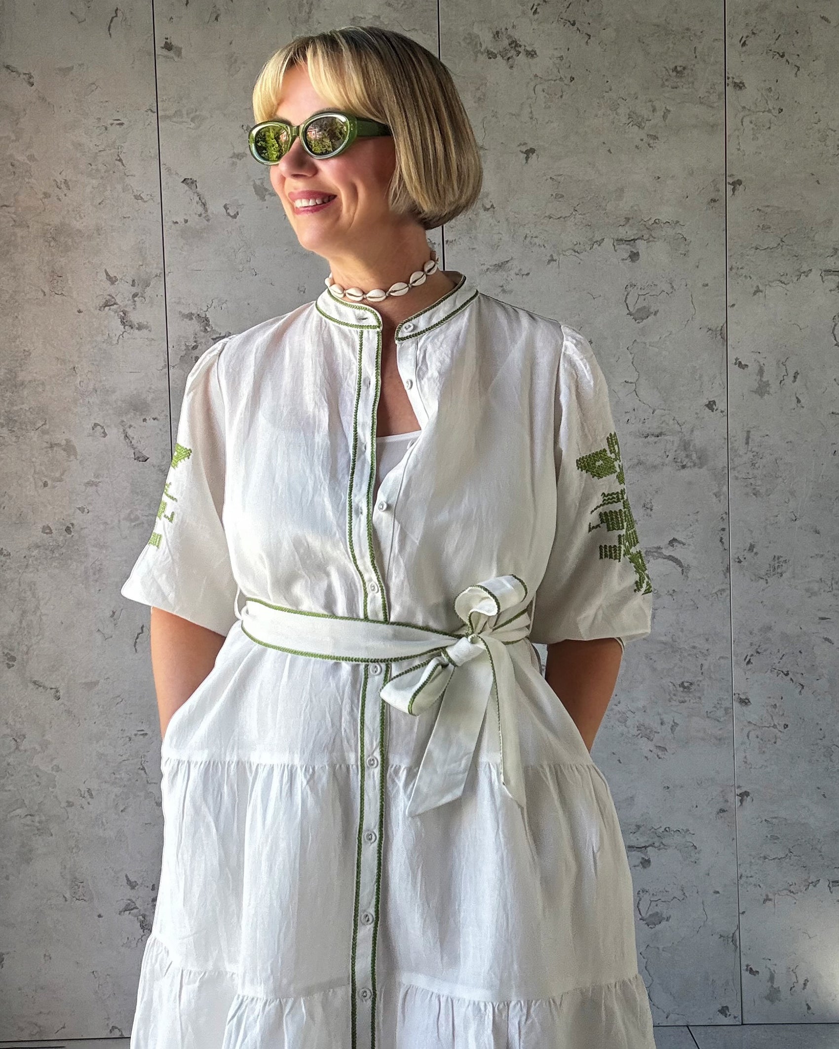 Woman wearing a white dress with green embroidery against a gray wall.