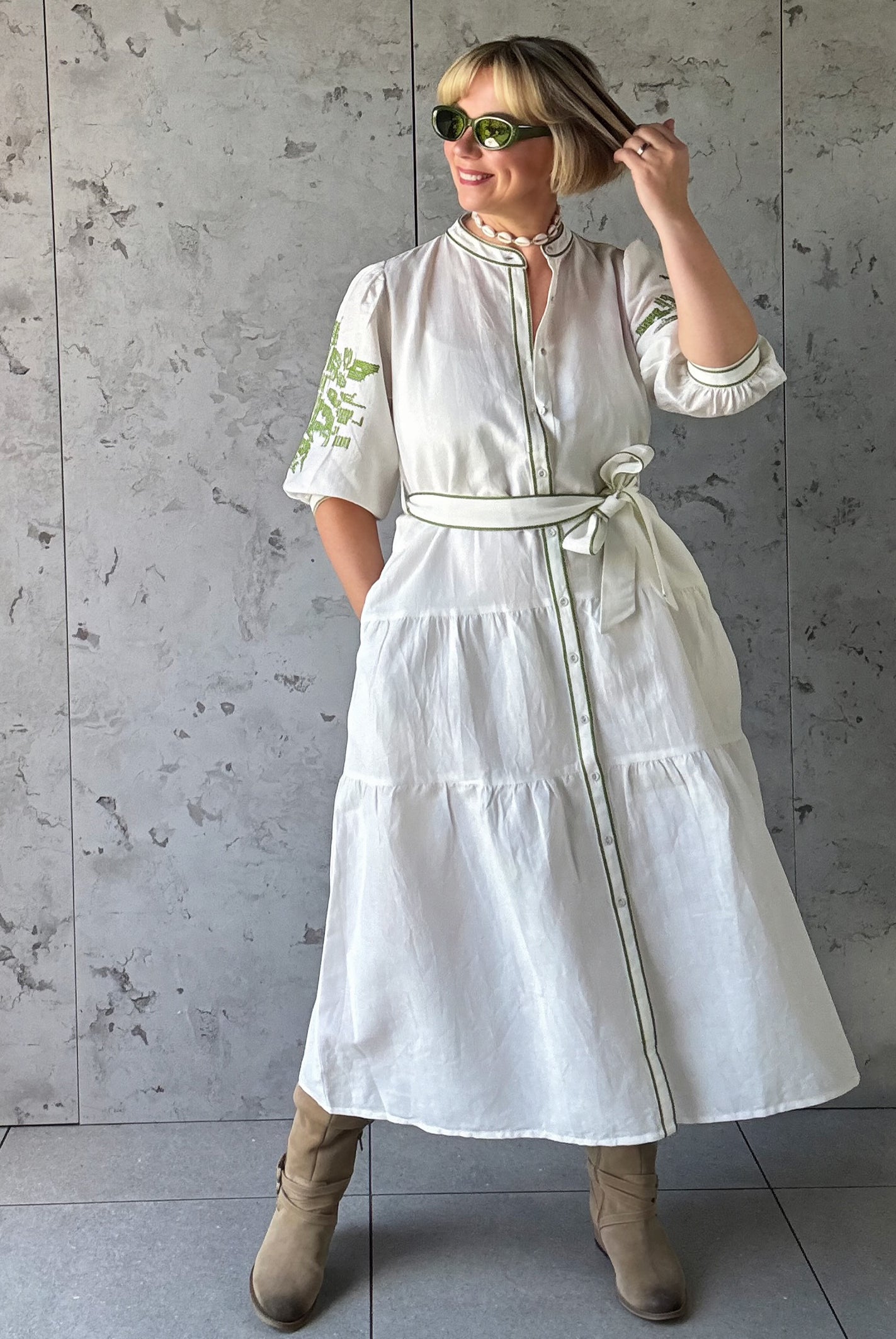 Woman wearing a white dress with green embroidery against a gray concrete wall.
