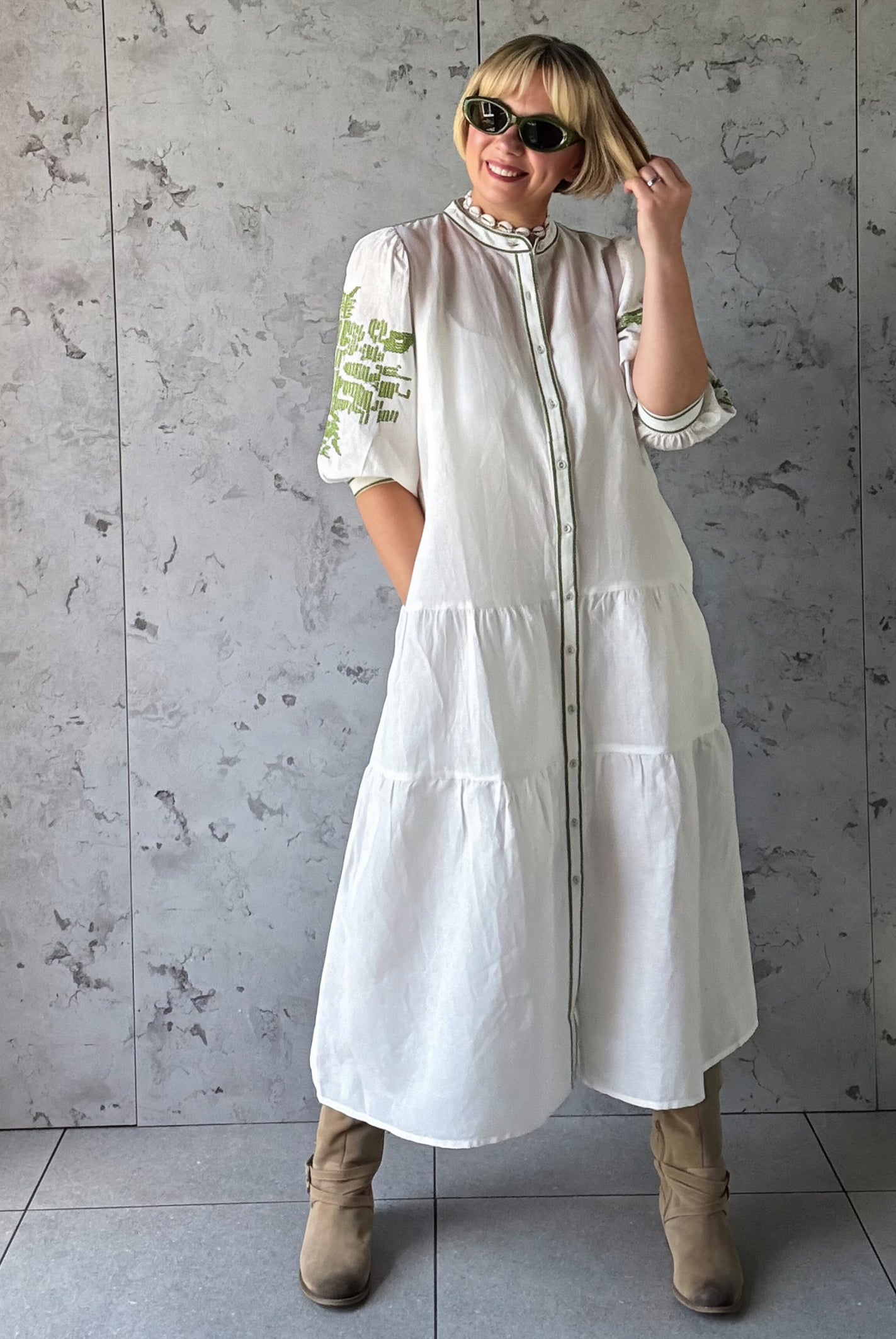 Woman wearing a white dress with green embroidery against a gray concrete wall.