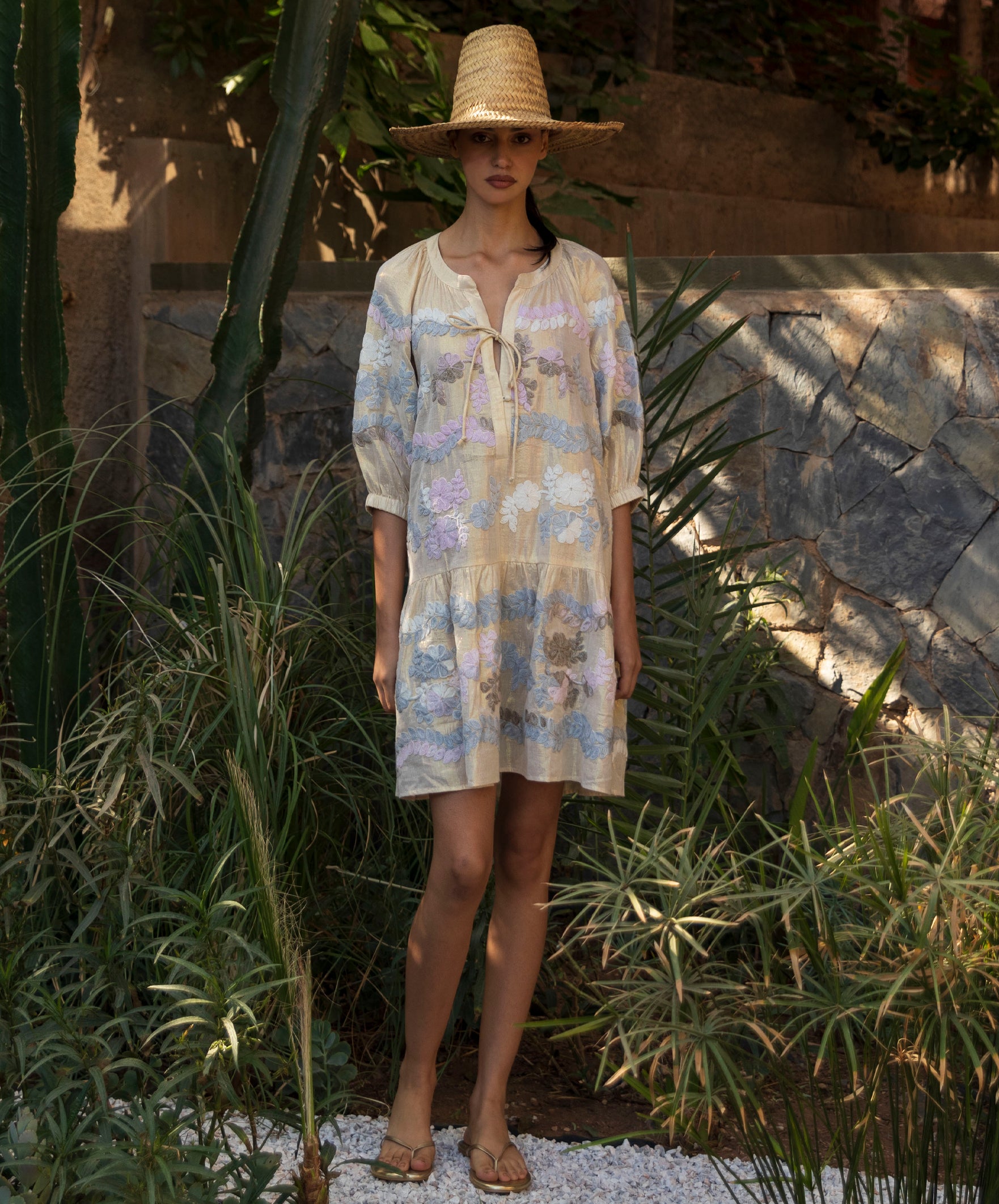 Woman wearing a floral embroidered gold Oleander dress and straw hat in a tropical garden setting