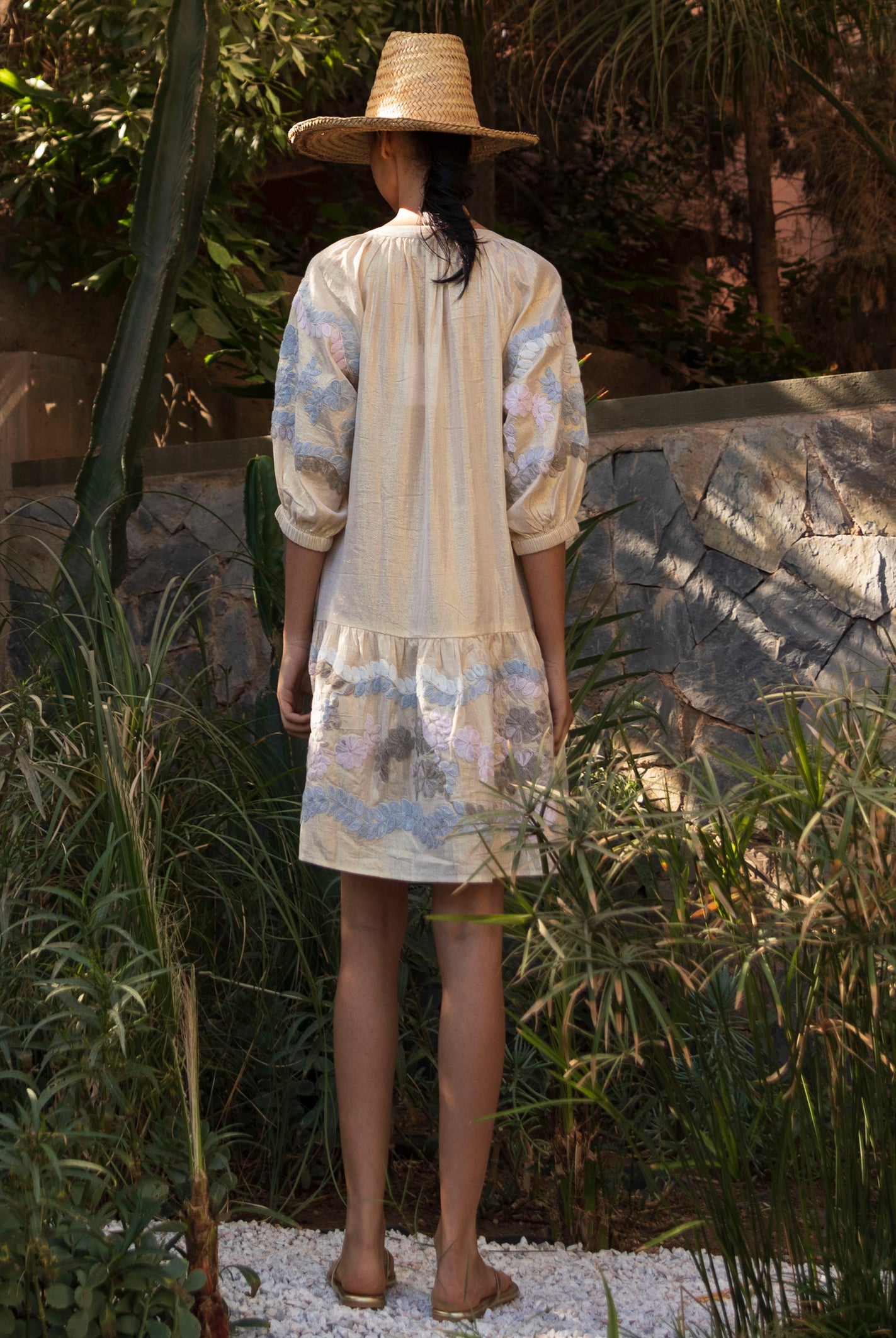 Back view of a woman wearing a floral embroidered gold Oleander dress and straw hat standing in a tropical garden setting