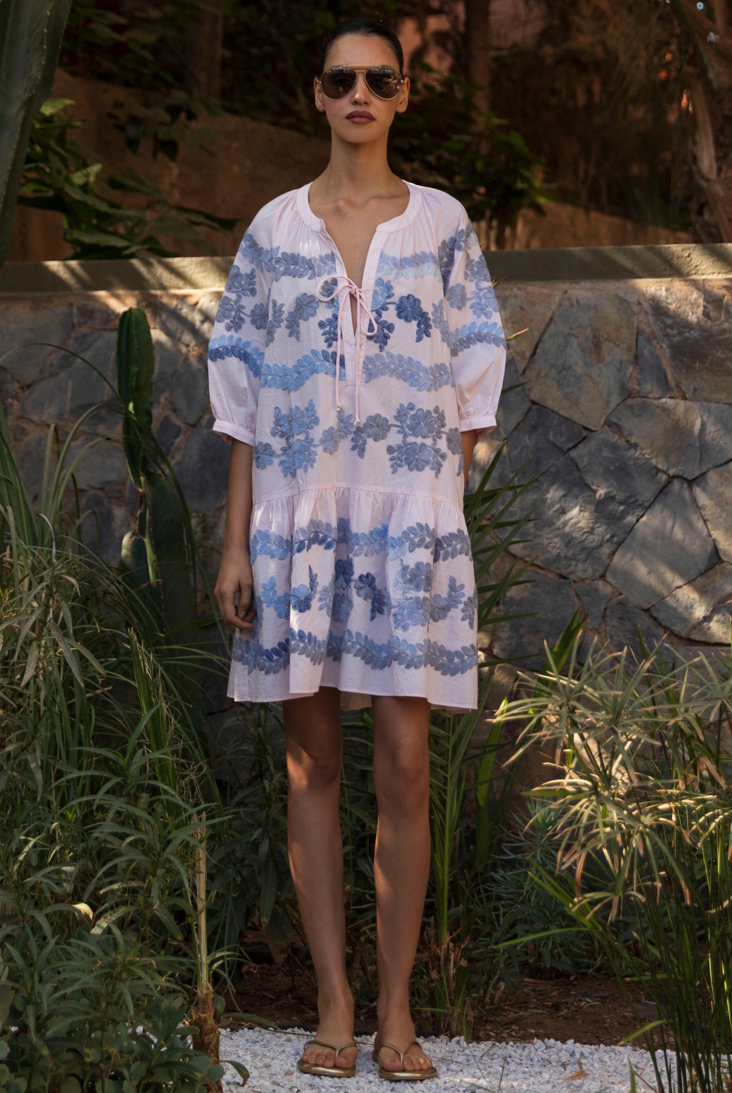 Woman wearing a light pink Oleander dress with blue floral embroidery standing in a tropical garden