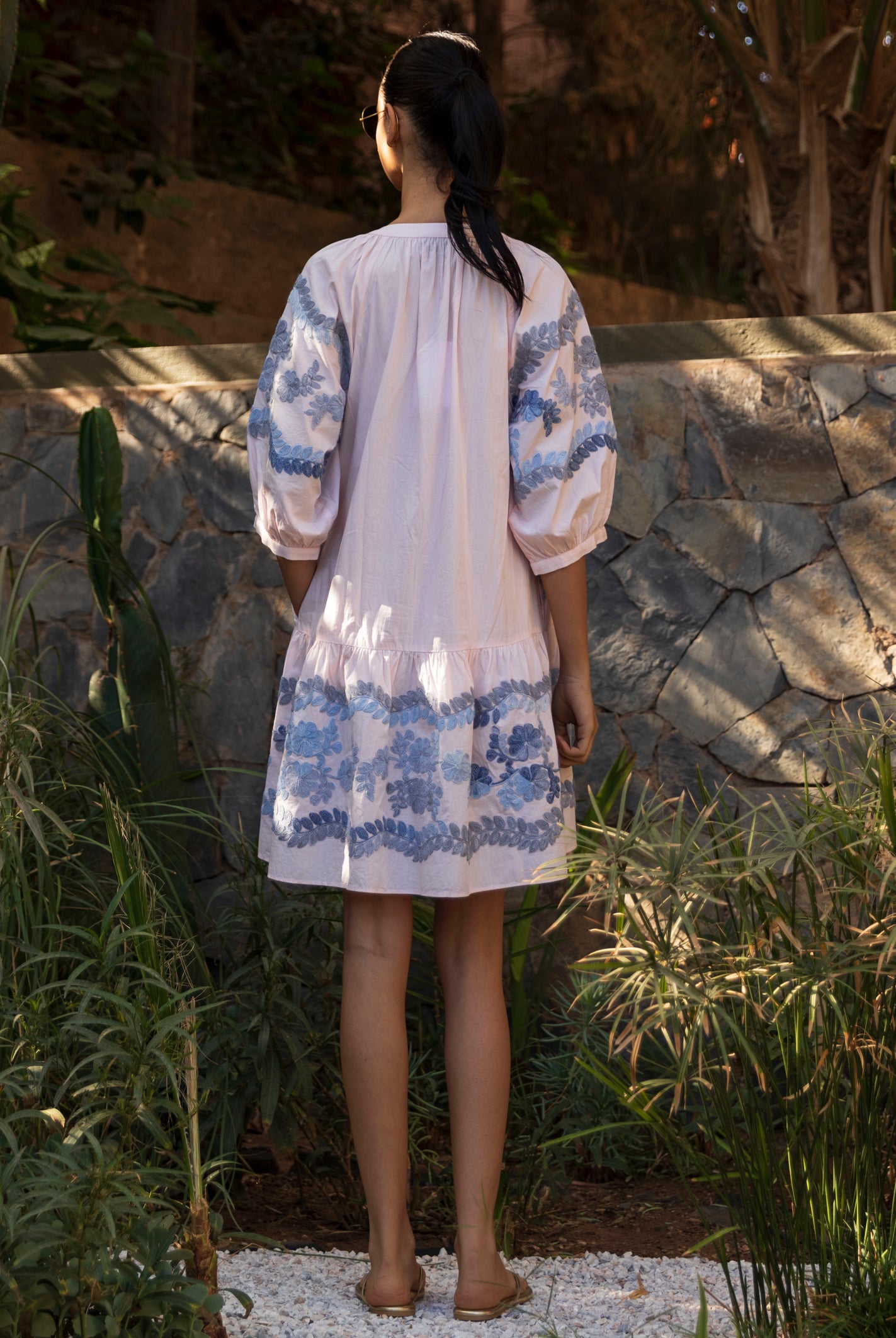 Back view of a woman in a light pink Oleander dress with blue floral embroidery standing in a tropical garden setting.