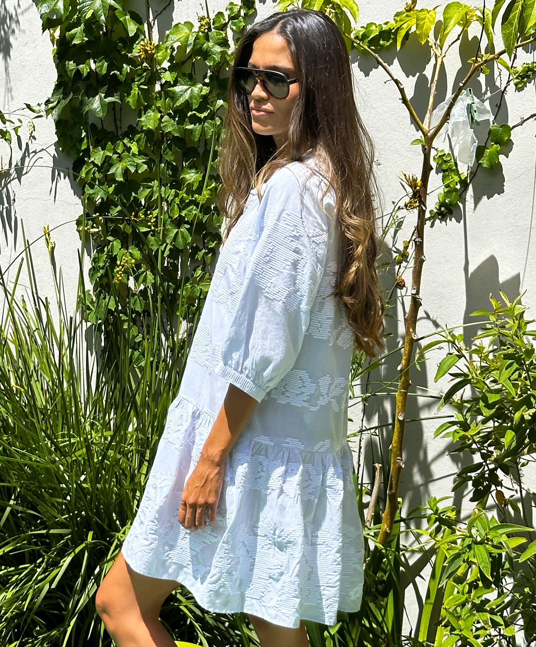 A model stood in a garden wearing sunglasses and a Rose and Rose pale blue cotton embroidered Palermo dress.
