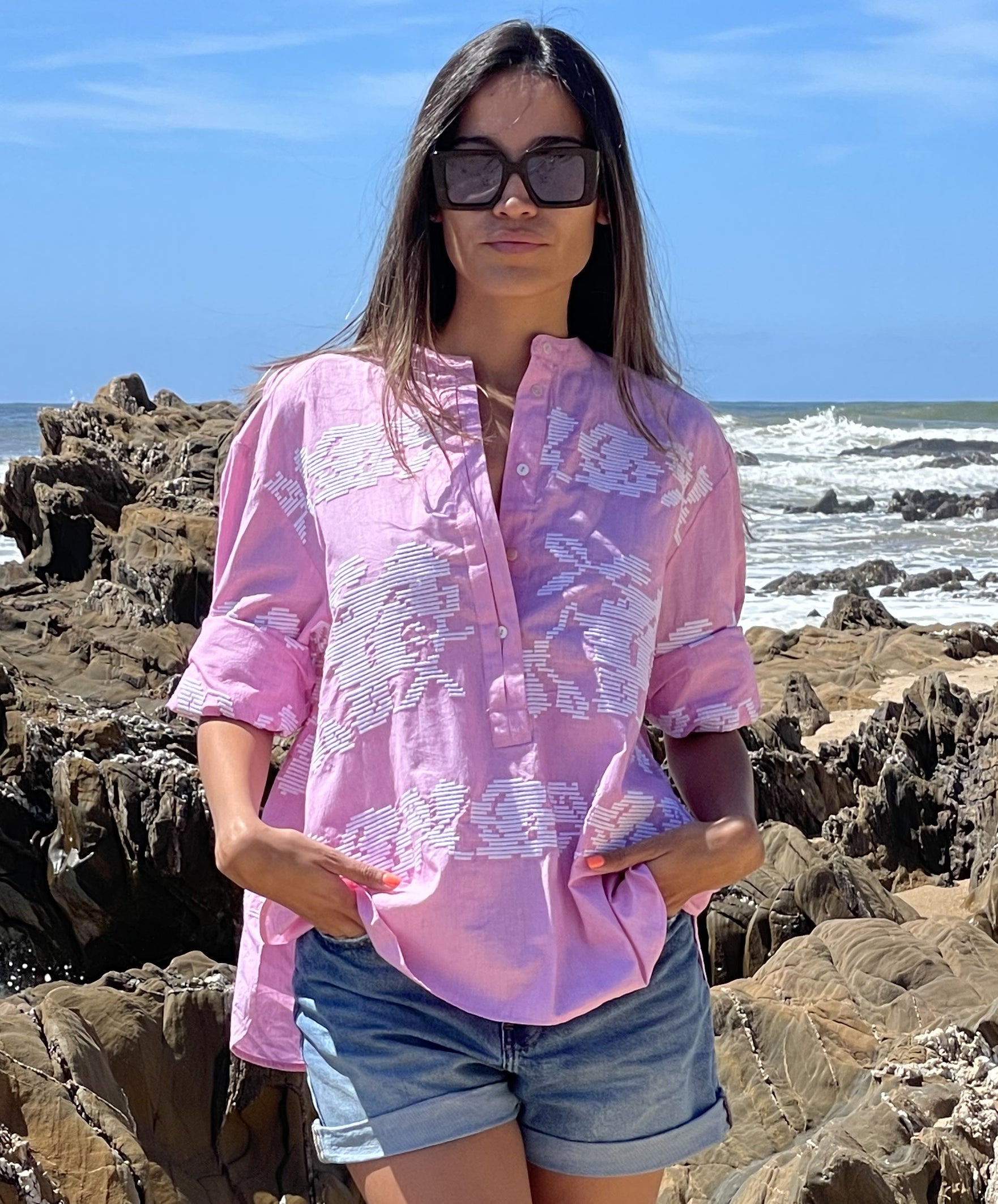 A model on a beach wearing a Rose and Rose pink Parma shirt, denim shorts and sunglasses. 