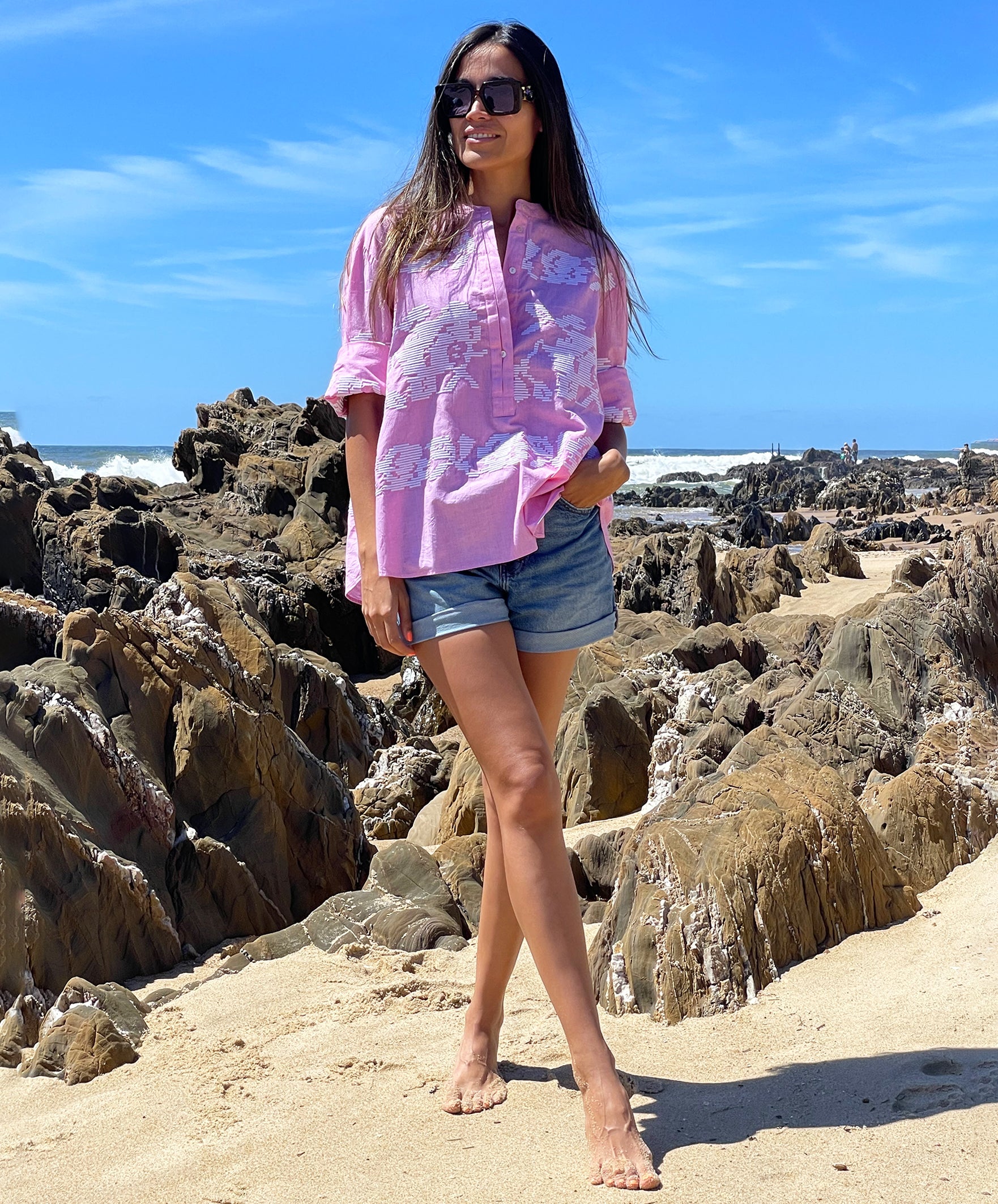 A model on a beach wearing a Rose and Rose pink Parma shirt, denim shorts and sunglasses. 
