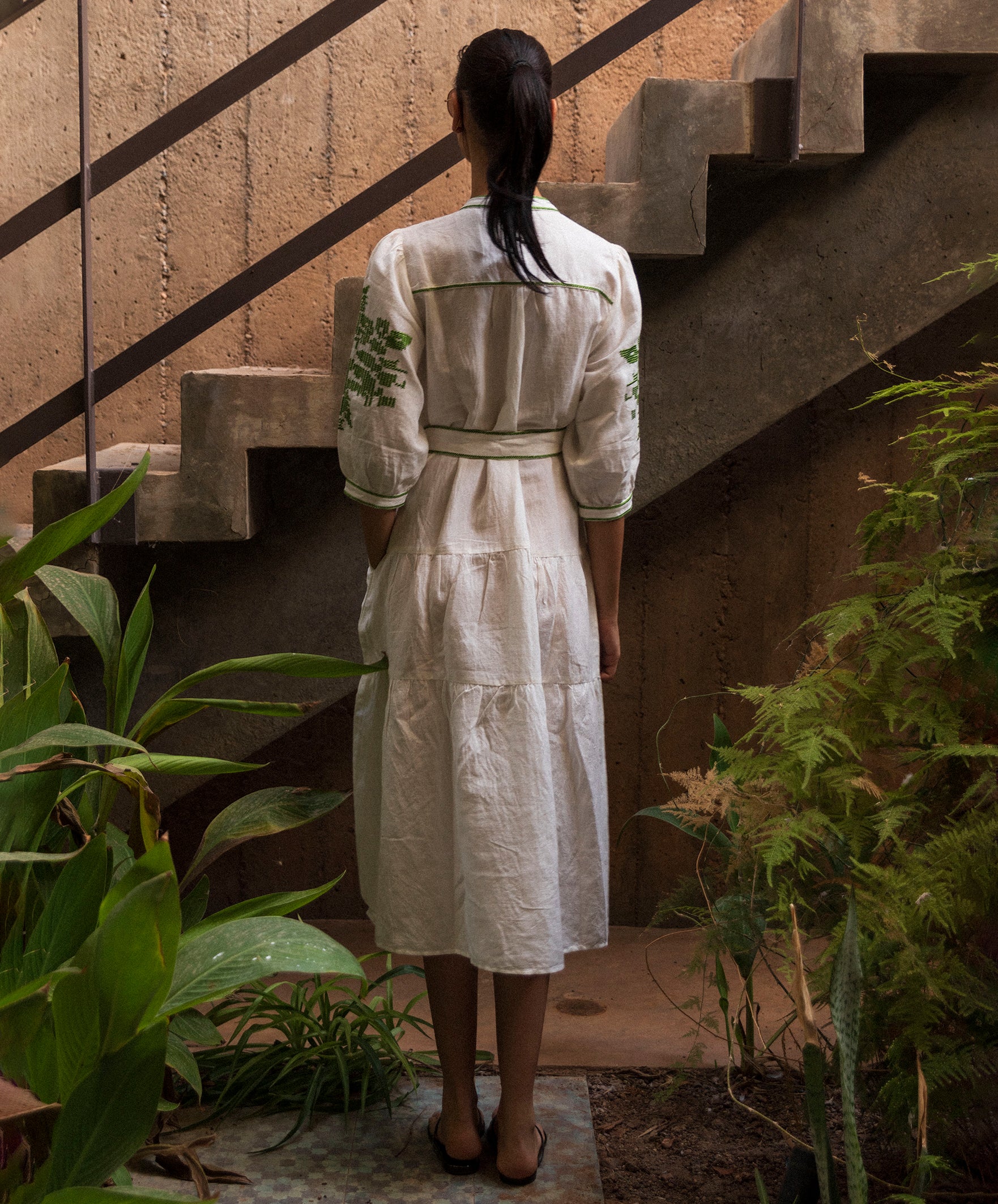 Woman in a white linen dress with olive green embroidery standing on a staircase with plants around