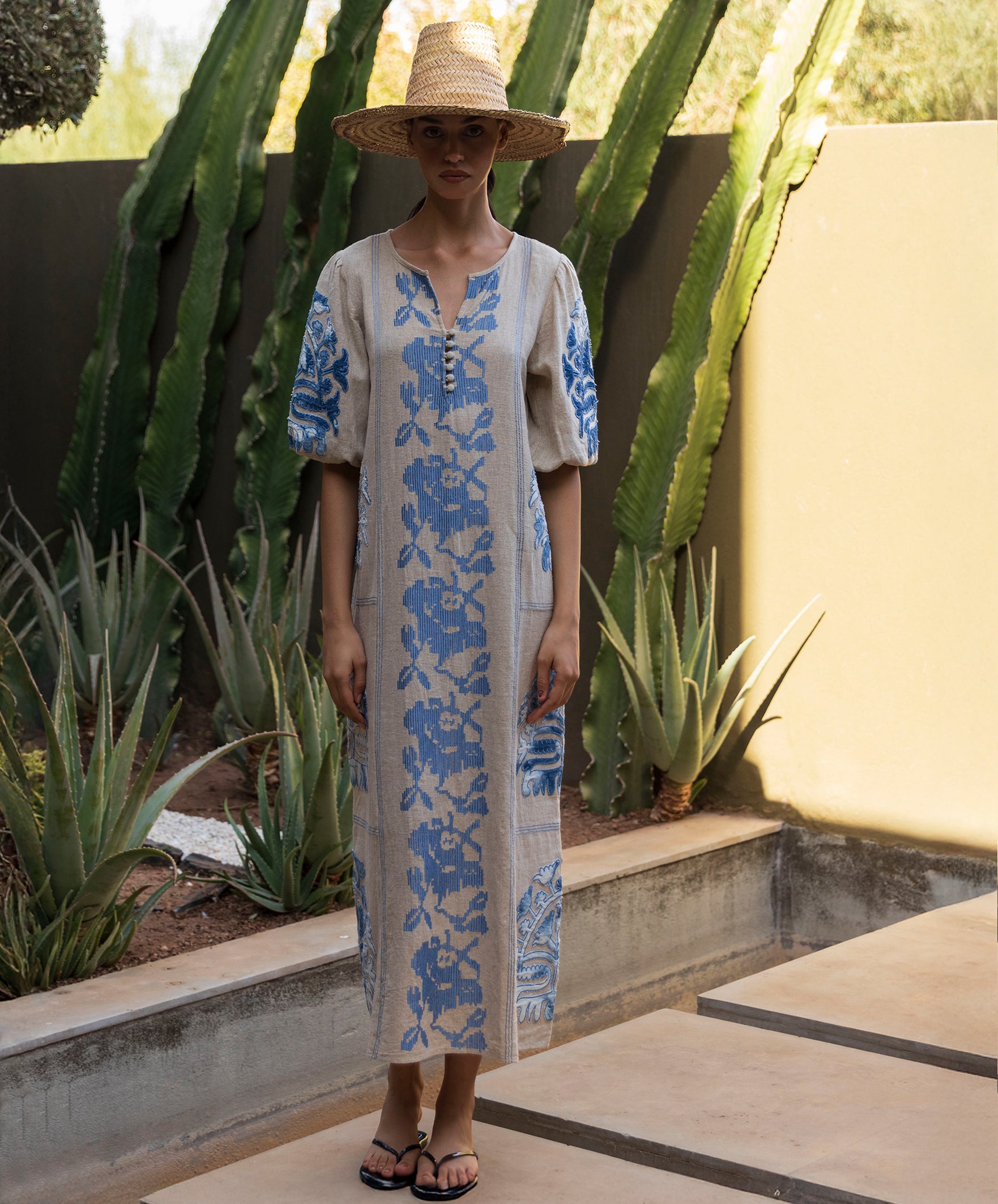 Woman wearing a long, Rose dress with blue embroidery and applique decoration in a cacti garden setting.