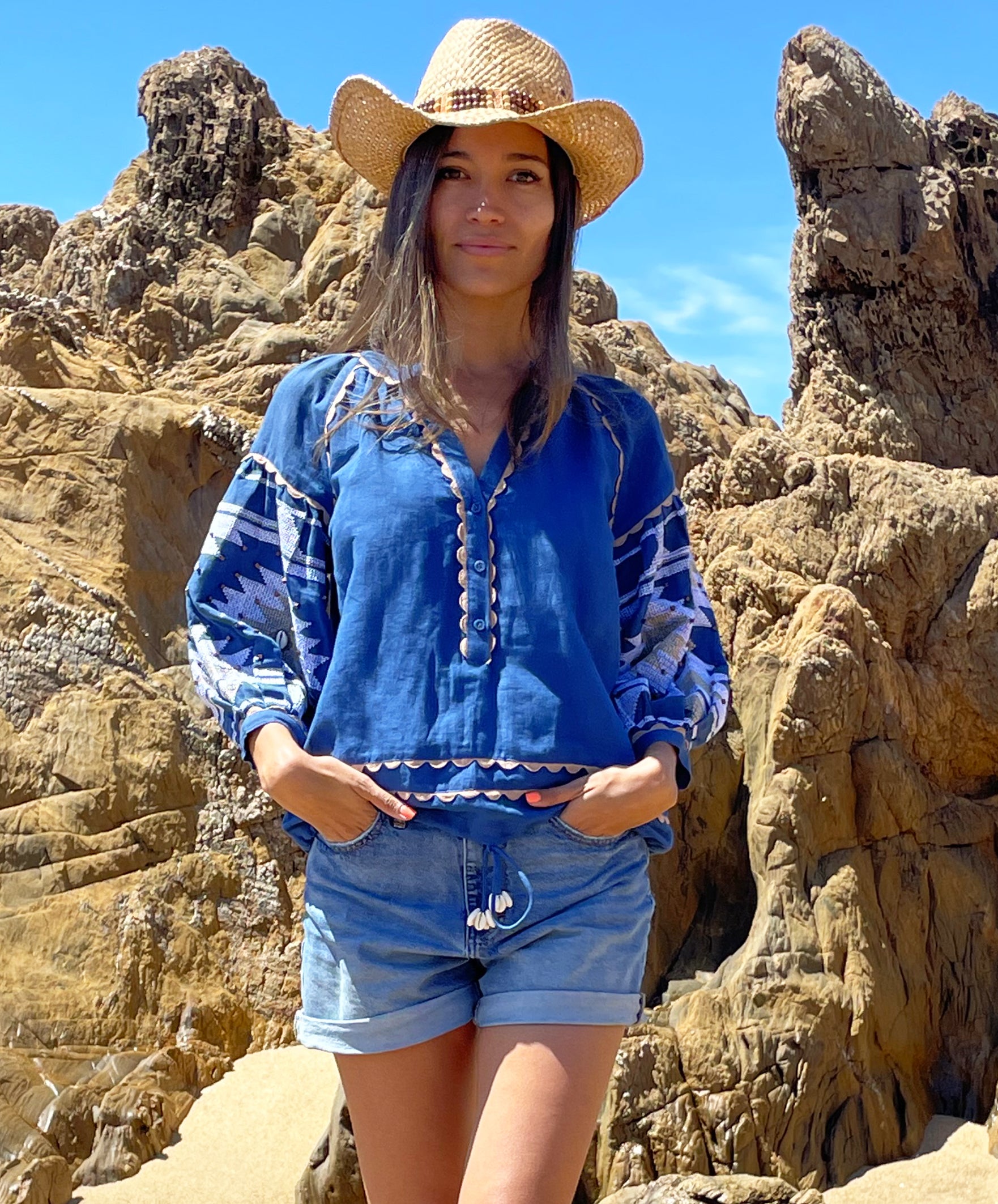 A model stood in front of beach rocks wearing a Rose and Rose blue embroidered San Marino top, denim shorts and a cowboy hat.