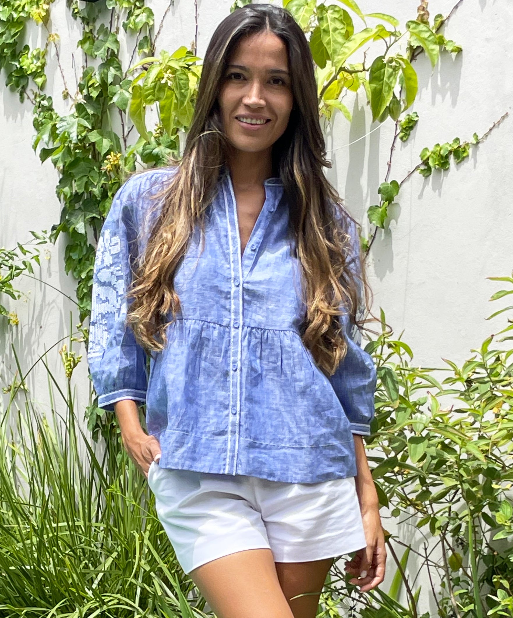 A mode stood in a garden wearing the blue chambray linen Rose and Rose Sardinia top and white shorts.
