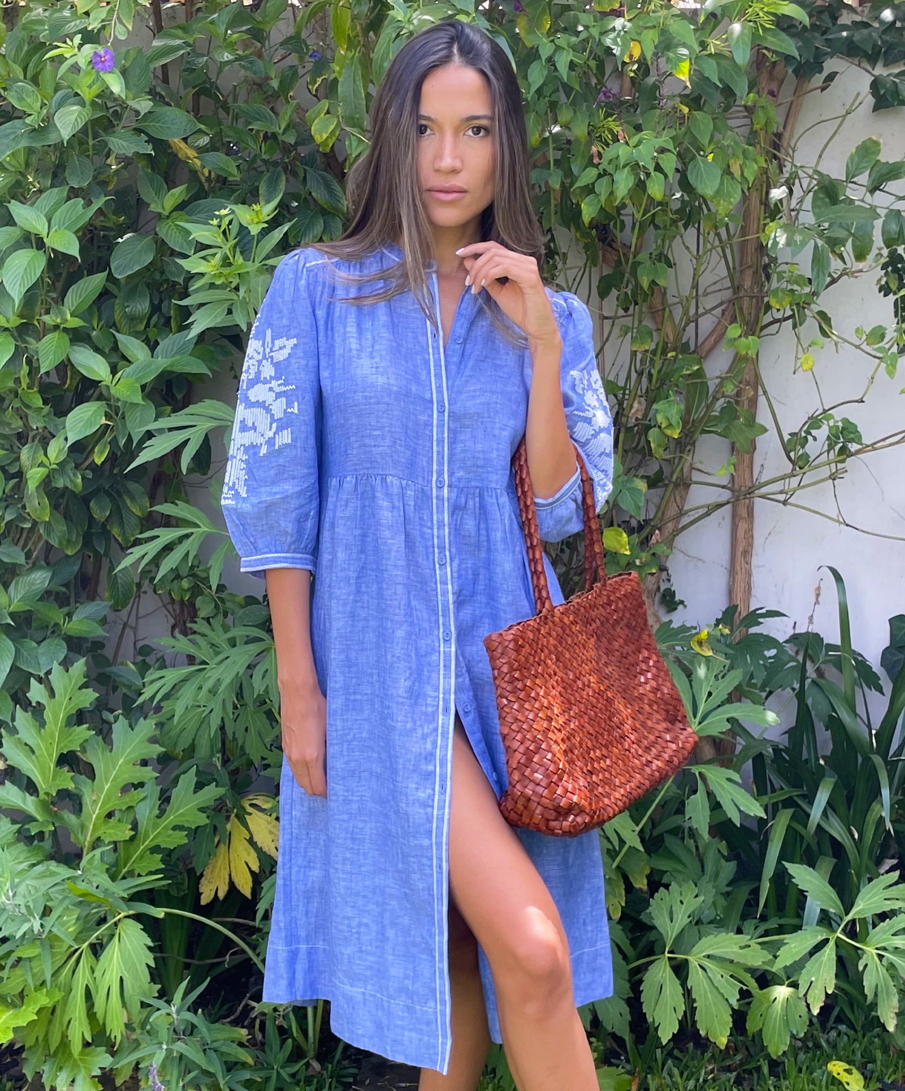 A model in a garden wearing a Rose and Rose blue linen Sicily dress and holding a Dragon Diffusion woven leather bag.