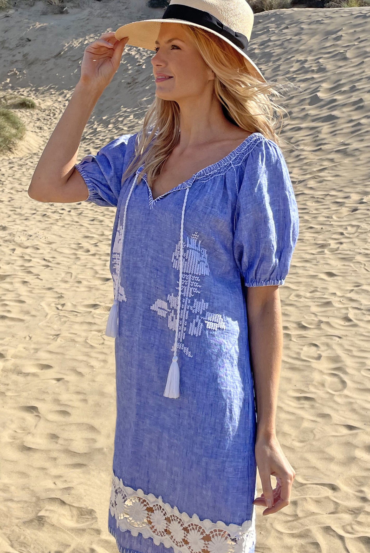 A model on a beach wearing the Rose and Rose Siracusa embroidered blue linen dress and an Anthony Peto Panama hat.