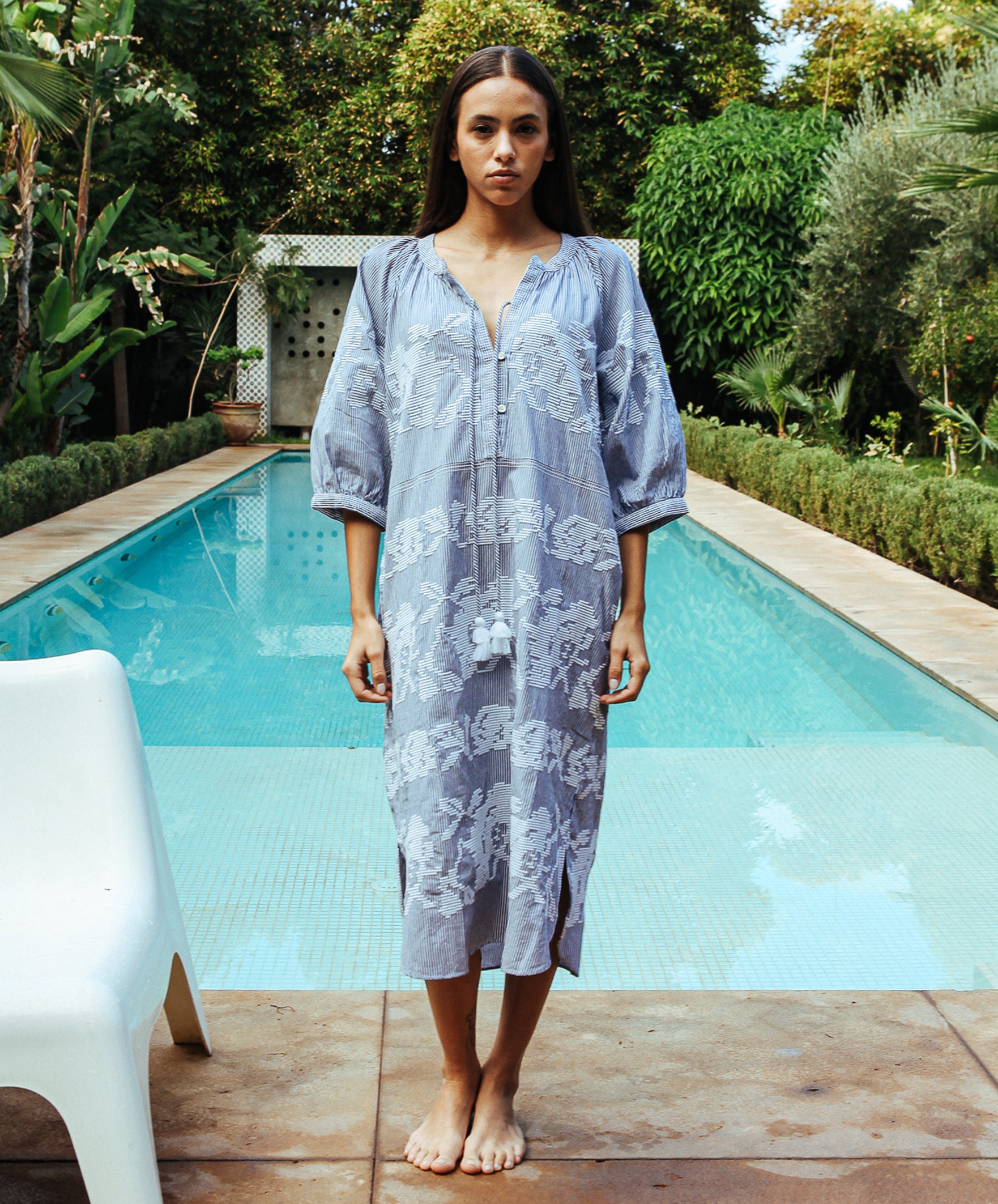 A model stood in front of a swimming pool wearing a Rose and Rose Snaresbrook striped cotton dress with white embroidery.