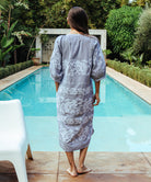 A back view of a model stood in front of a swimming pool wearing a Rose and Rose Snaresbrook striped cotton dress with white embroidery.
