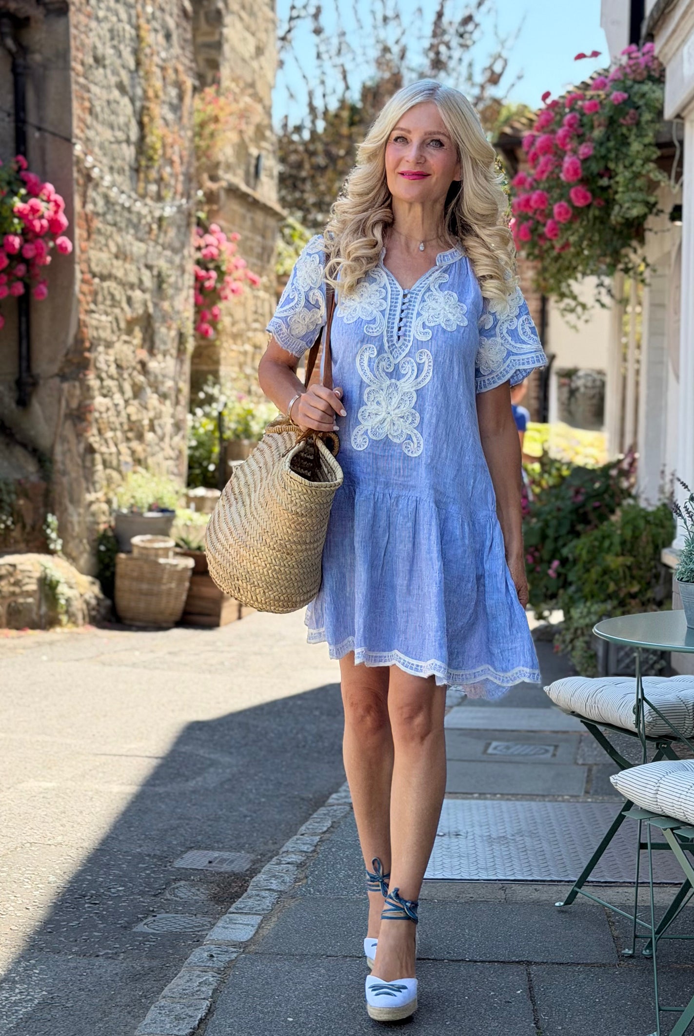 Influencer @seenonsarah walking down a pretty street wearing the Amaranta striped blue shirt dress with white floral embroidery, carrying a large woven straw tote bag.