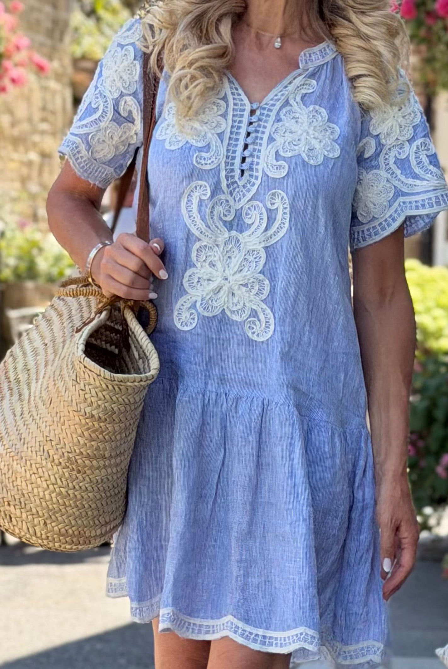 A woman wearing a striped blue short-sleeve Amaranta dress with white floral embroidery, holding a large woven straw tote bag in an outdoor garden setting.