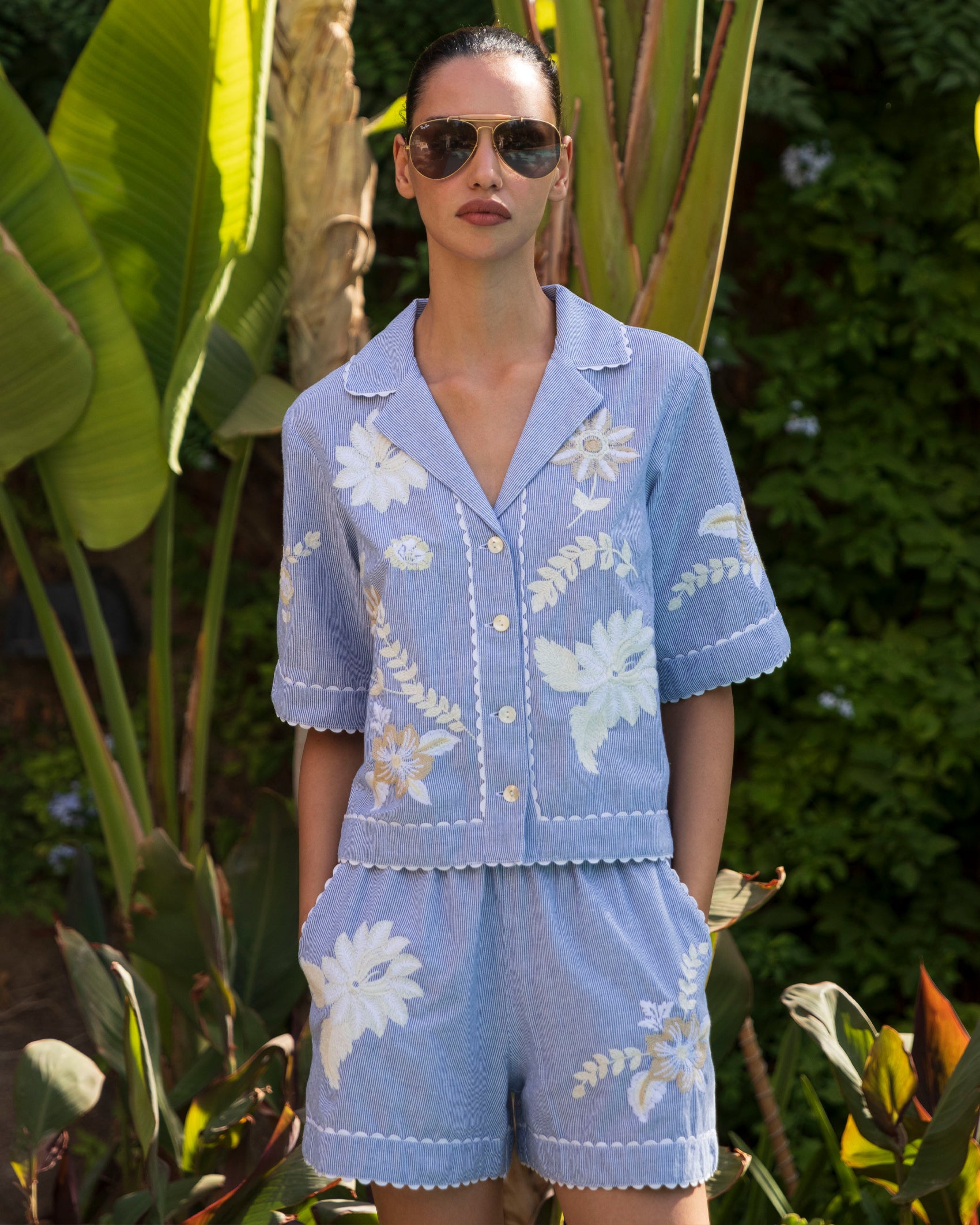 Woman wearing a blue striped embroidered blouse and shorts set with sunglasses in a garden setting