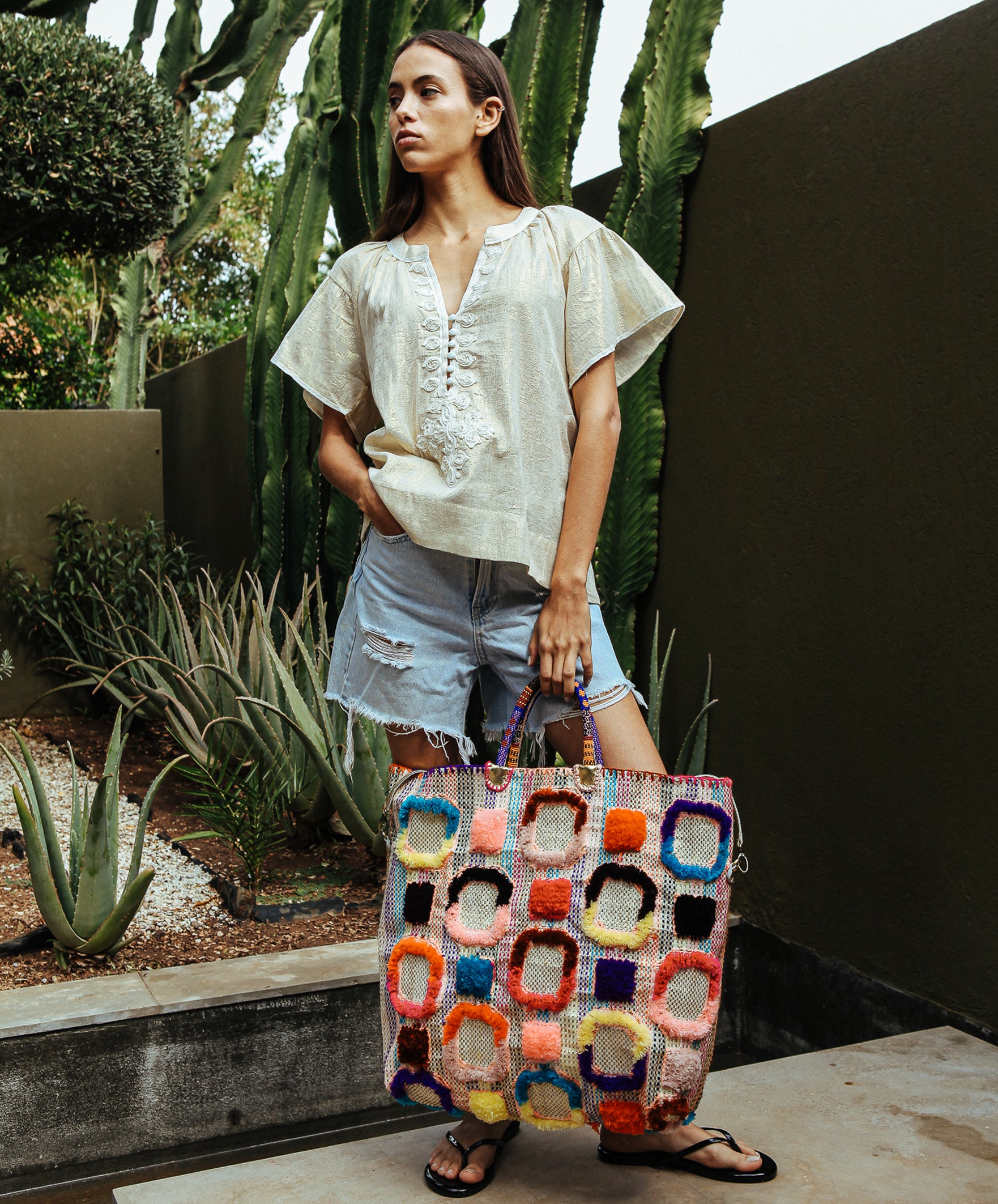 A model stood in a cacti garden wearing a Rose and Rose Upminster gold top, denim shorts and a Foodbag Foudation bag.