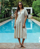 A model stood in front of a swimming pool wearing a Rose and Rose Uxbridge gold dress with white passementerie decoration.