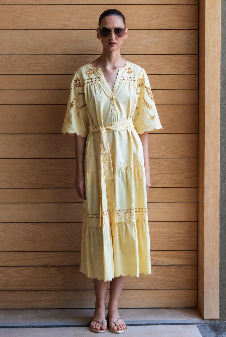 Woman wearing a light yellow embroidered dress and straw hat against a wooden wall.