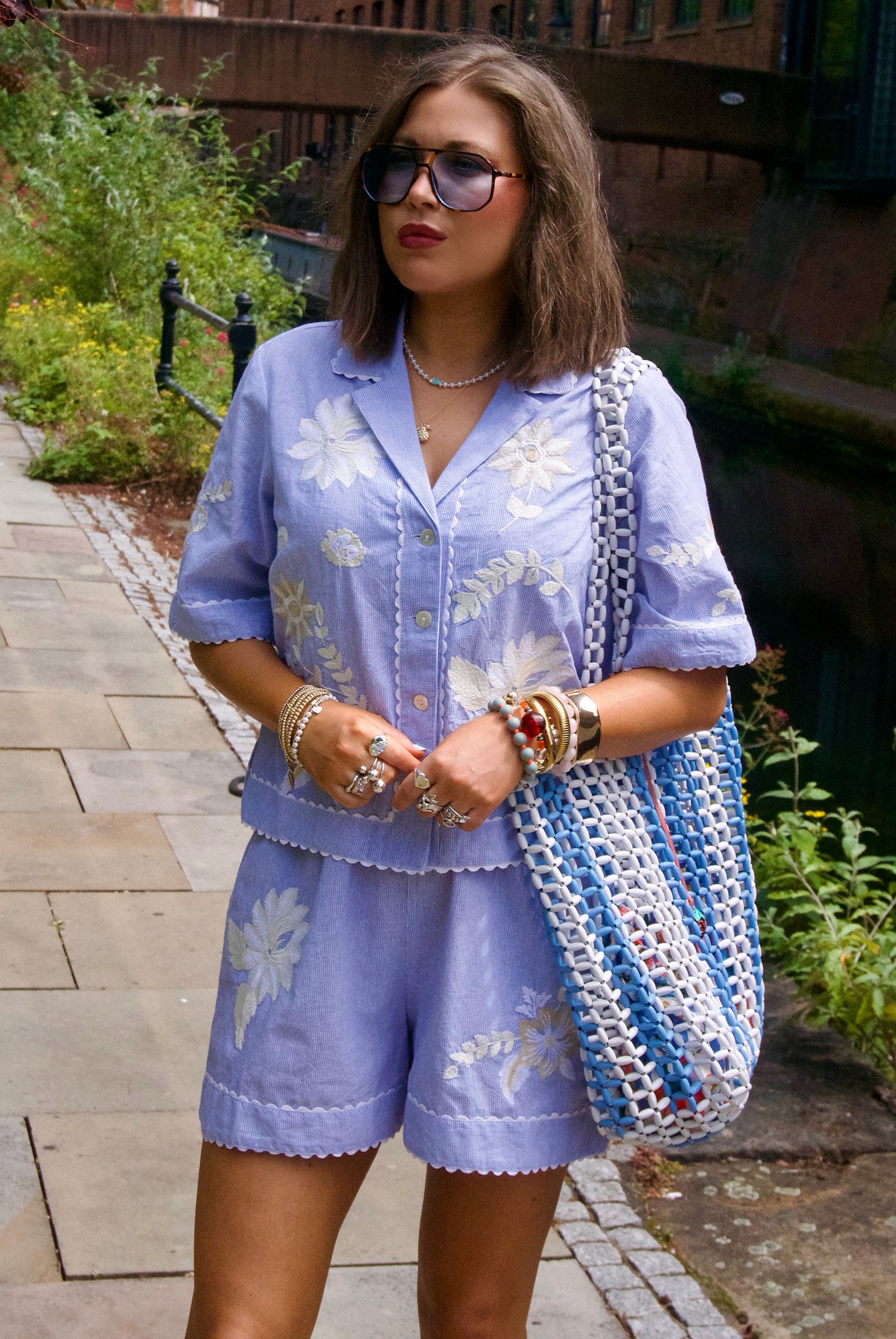 Influencer @irenadworld wearing a light blue striped short-sleeve button-up shirt and matching shorts set with white floral embroidery, styled with a blue and white tote bag, sunglasses, and bracelets, standing outdoors on a stone path.