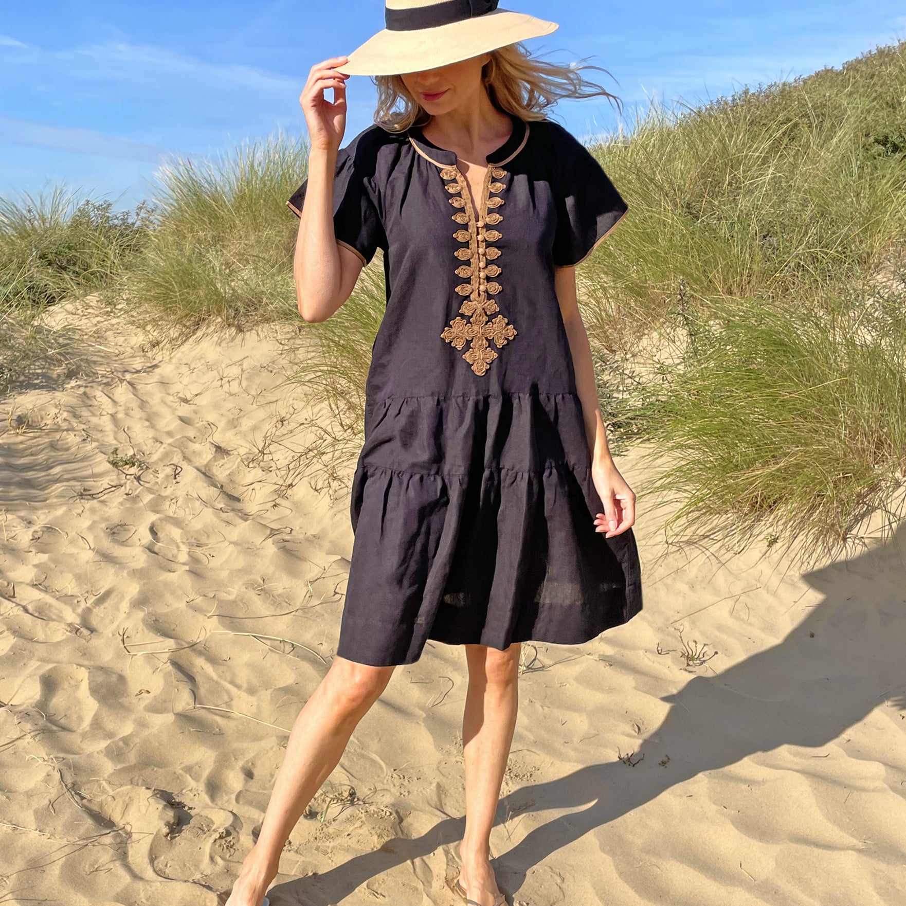 A model on the beach wearing a Rose and Rose black Ischia dress with passementerie decoration and an Anthony Peto Panama hat. 