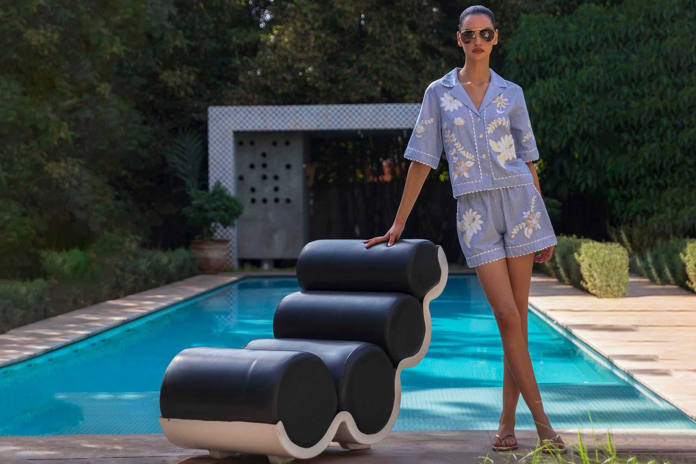 Woman in a blue striped floral embroidered shirt and shorts outfit standing next to modern outdoor furniture by a pool.