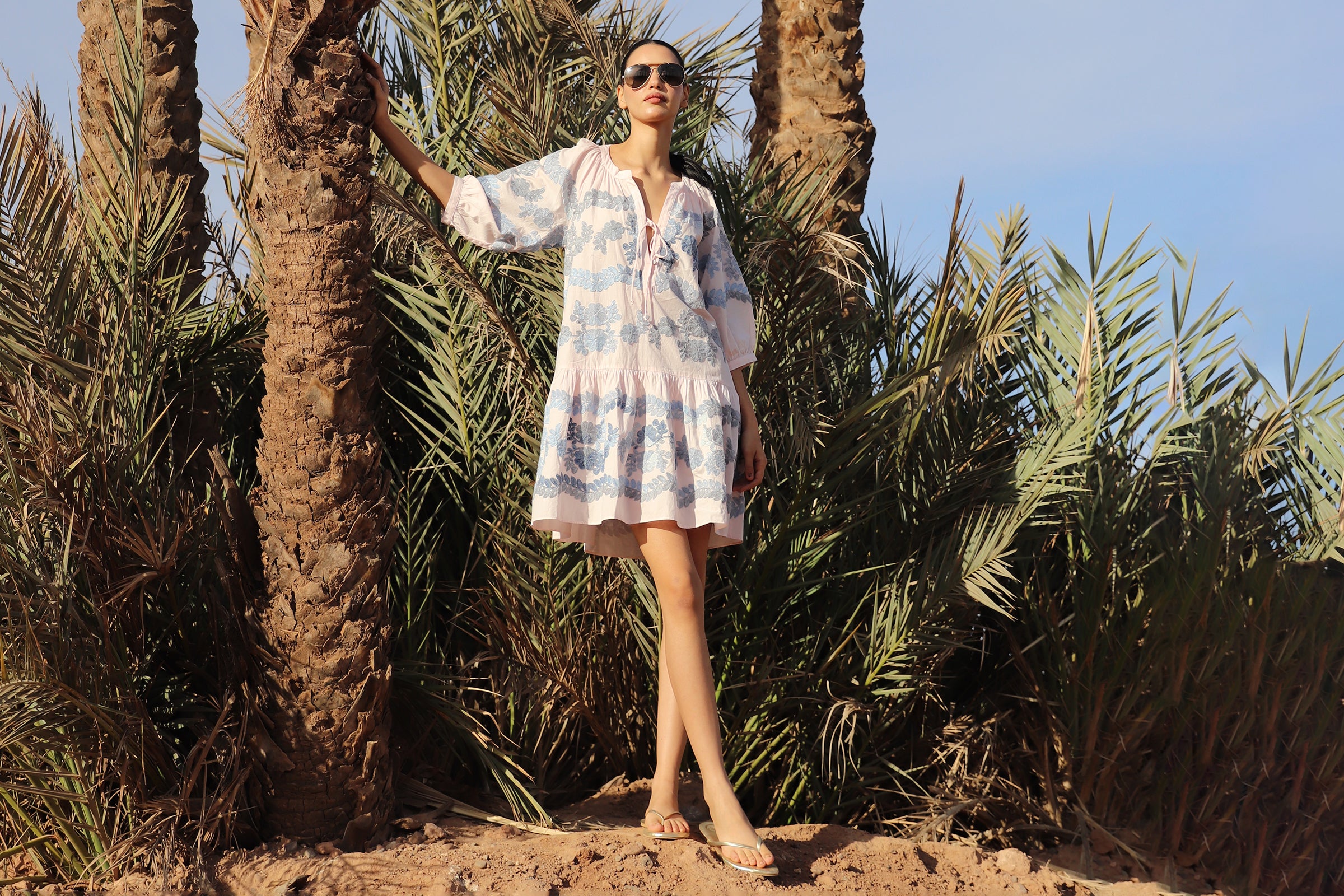 Woman in a pale pink dress with floral embroidery standing among palm trees