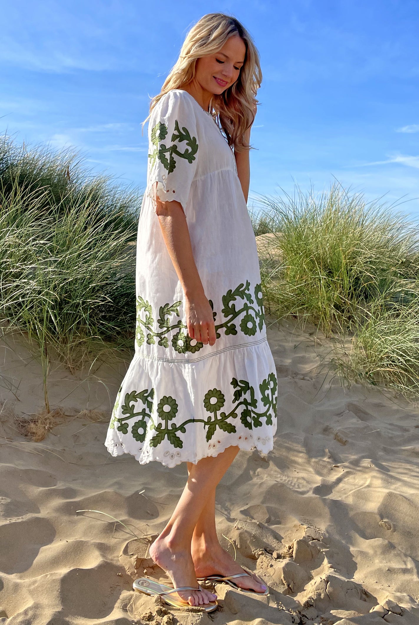 A side view of a model on a beach wearing the Rose and Rose Agrigento dress in white linen with olive applique.