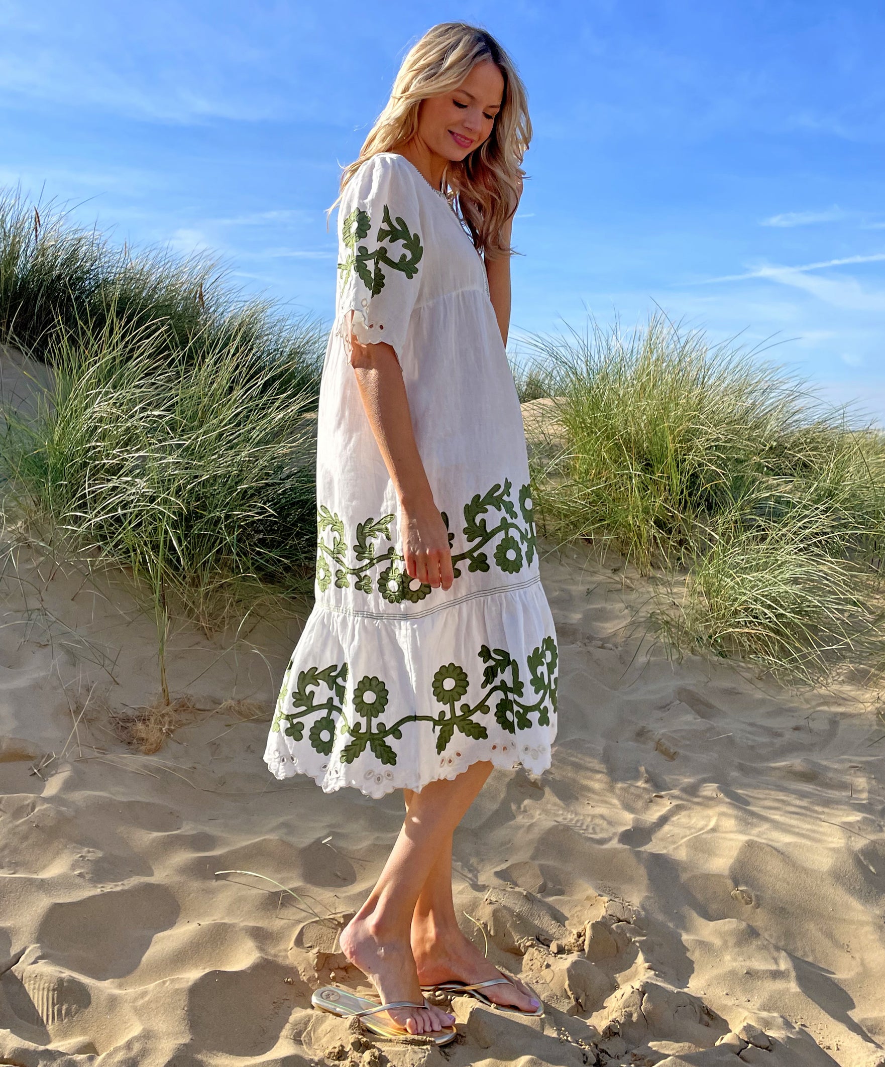A side view of a model on a beach wearing the Rose and Rose Agrigento dress in white linen with olive applique.