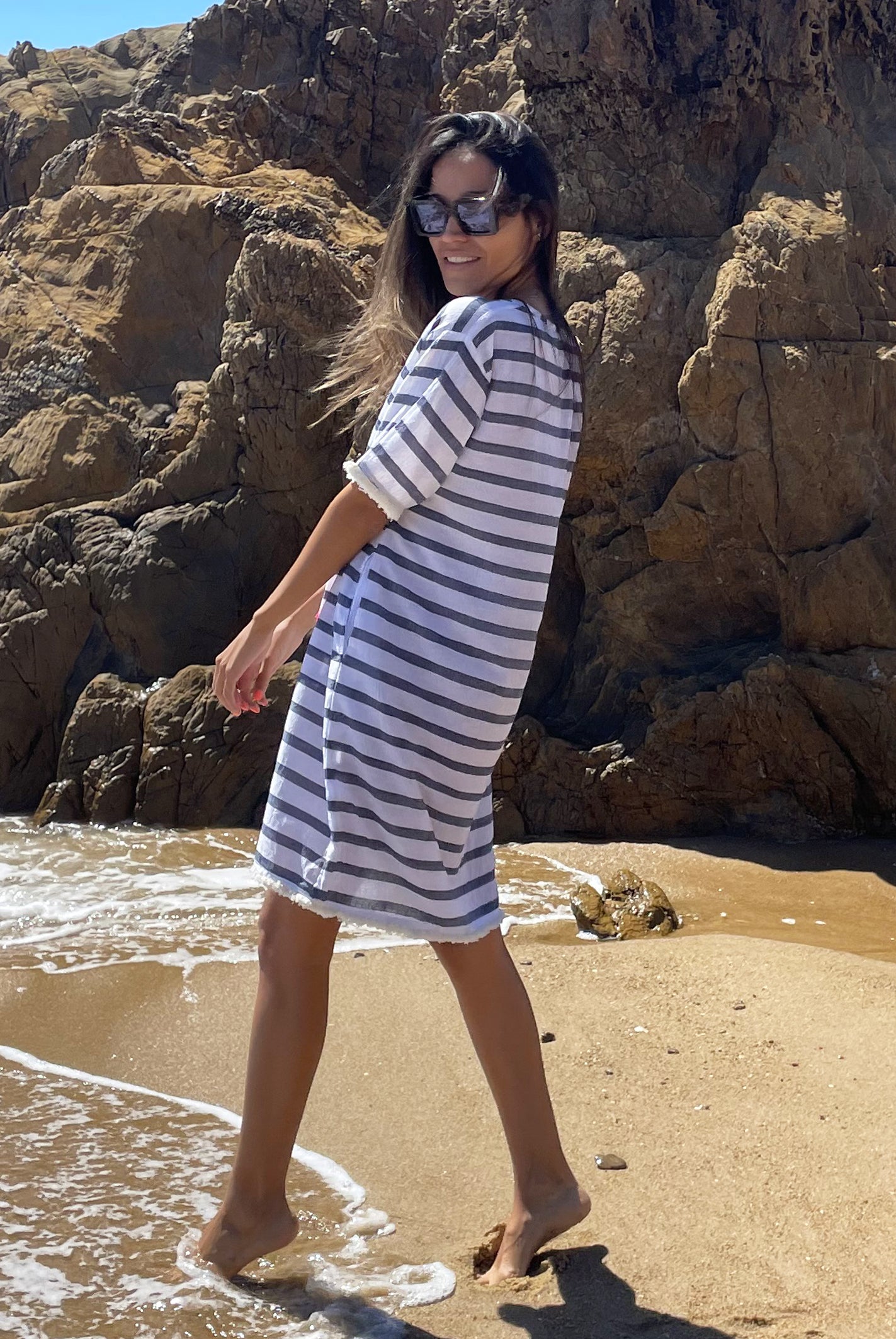 A back view of a model standing on the beach wearing a Rosebud by Rose and Rose Aix beach dress and sunglasses.
