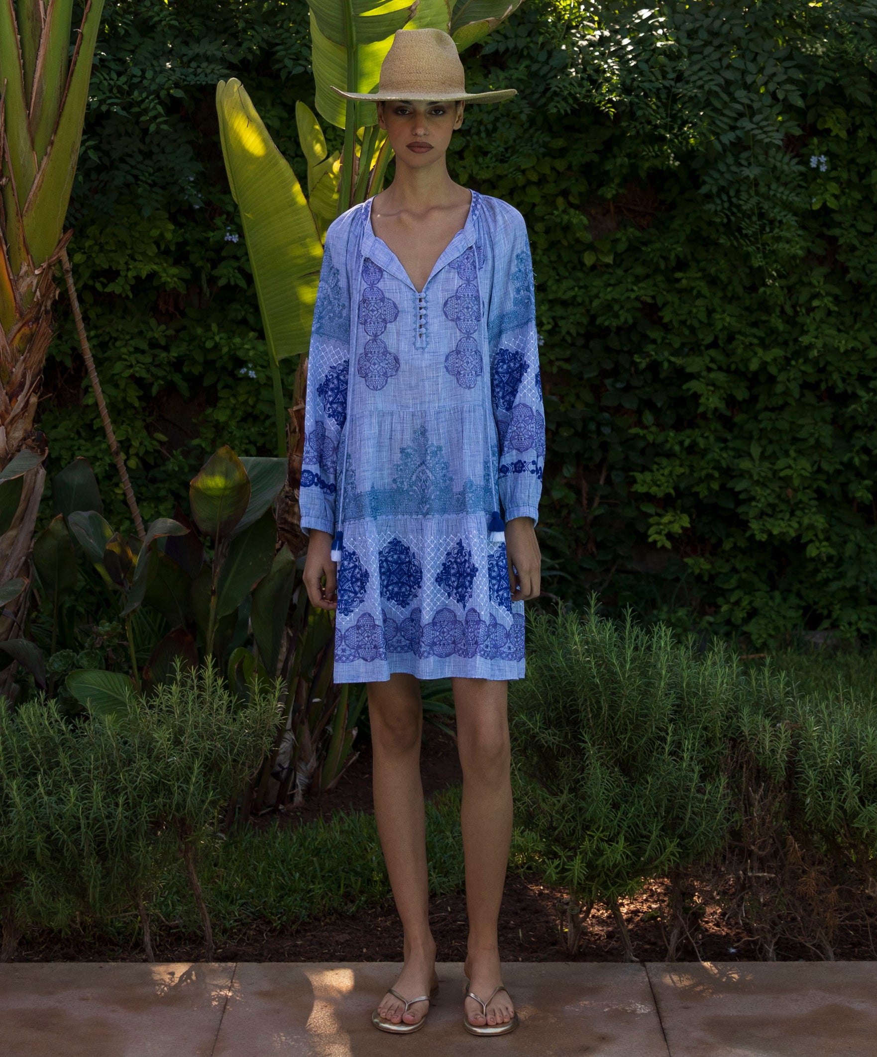 Woman wearing a blue floral embroidered Alder dress and straw hat in a tropical garden setting
