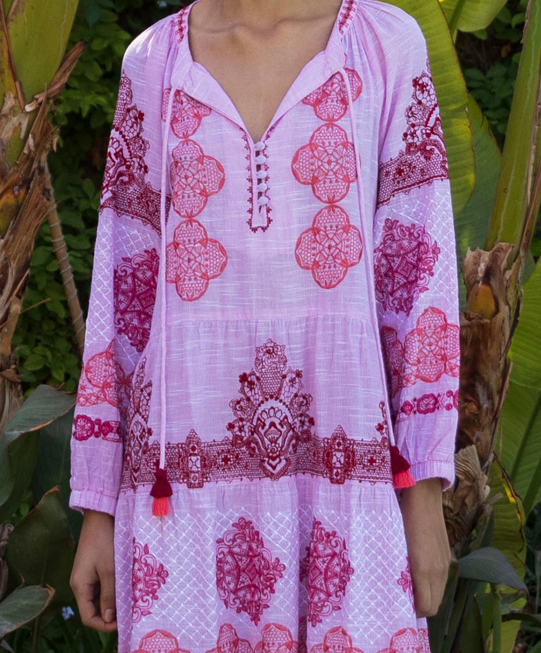 Close up of model wearing a pink and red embroidered Alder dress with tassels in a tropical setting.