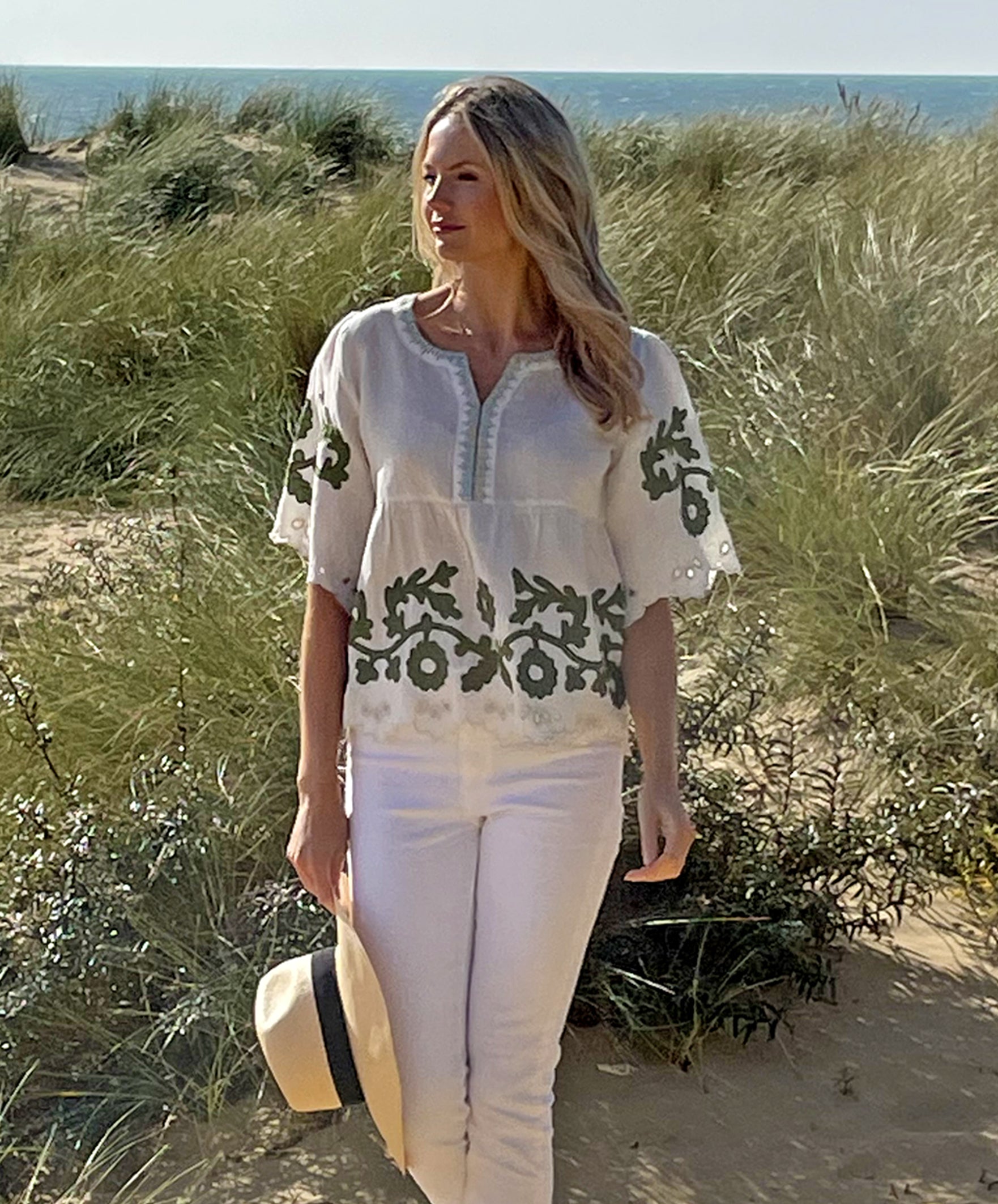 A model on a beach wearing the Rose and Rose Alessio top in white linen with olive applique and holding an Anthony Peto Panama hat. 