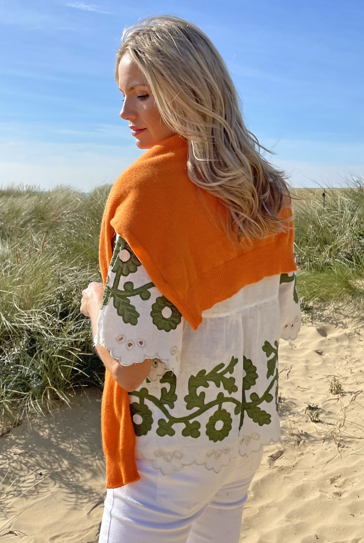 A back view of a model on a beach wearing the Rose and Rose Alessio top in white linen with olive applique and a Jumper 1234 jumper over her shoulders.