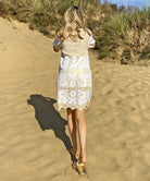 A back view of a model on a beach wearing the Rose and Rose Amalfi dress in gold lurex with white applique.