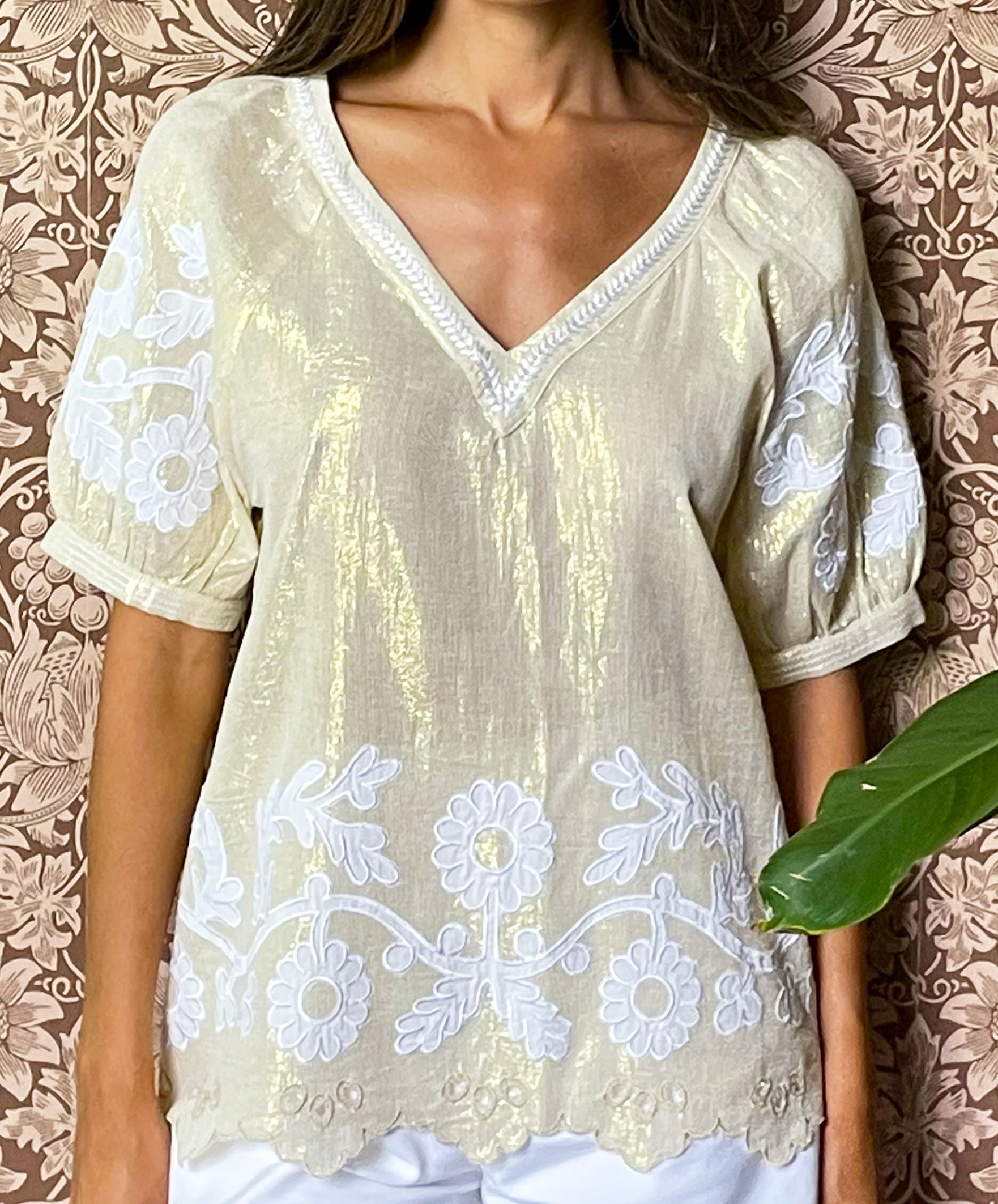 A close up of model stood in front of floral wallpaper wearing the Rose and Rose gold lurex Anacapri top with white applique. 