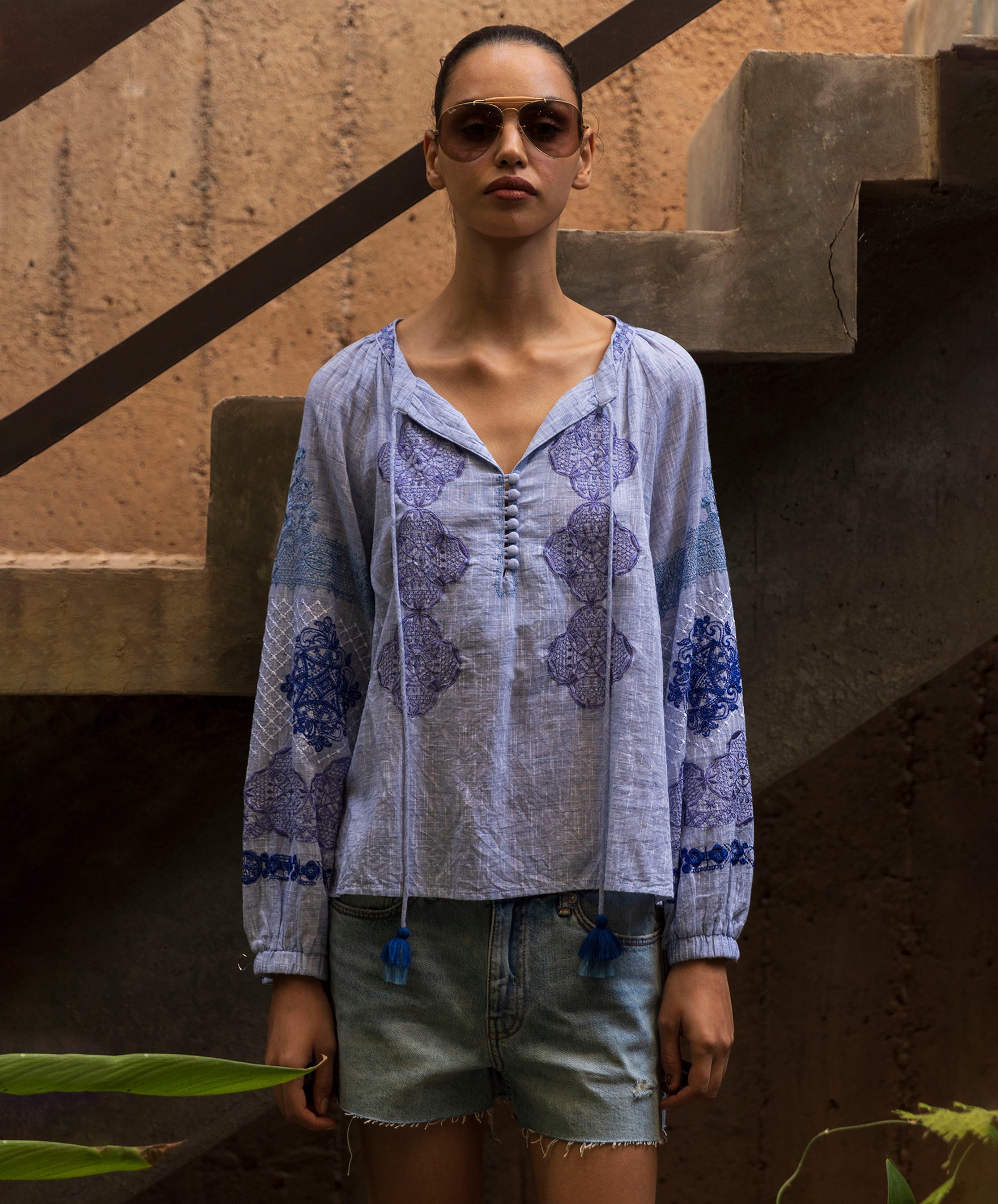 Woman wearing a floral embroidered blue Ash top and denim shorts standing against a textured wall.