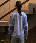 Woman wearing a blue melange floral embroidered Ash top standing in front of a textured wall.