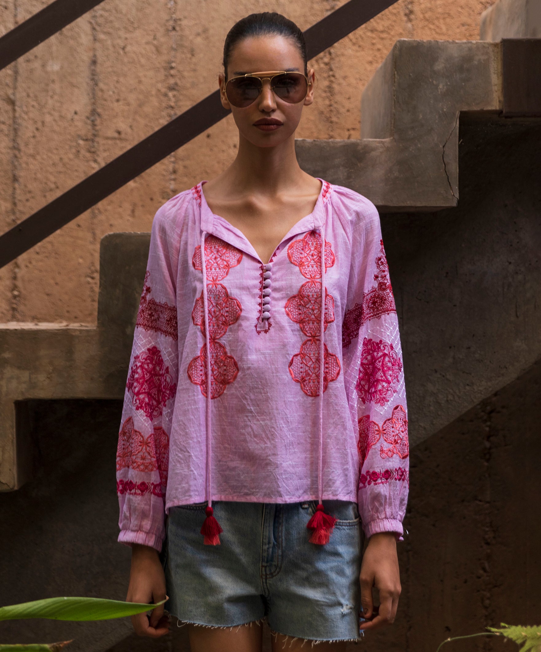 Woman wearing a pink floral embroidered Ash top and denim shorts, standing against a textured wall.