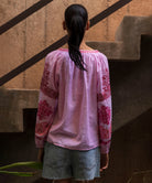 Woman wearing a pink blouse with floral embroidery standing in front of a textured wall.