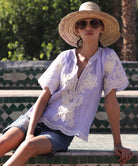 Woman sat on steps wearing a blue striped embroidered Azalea top and straw hat
