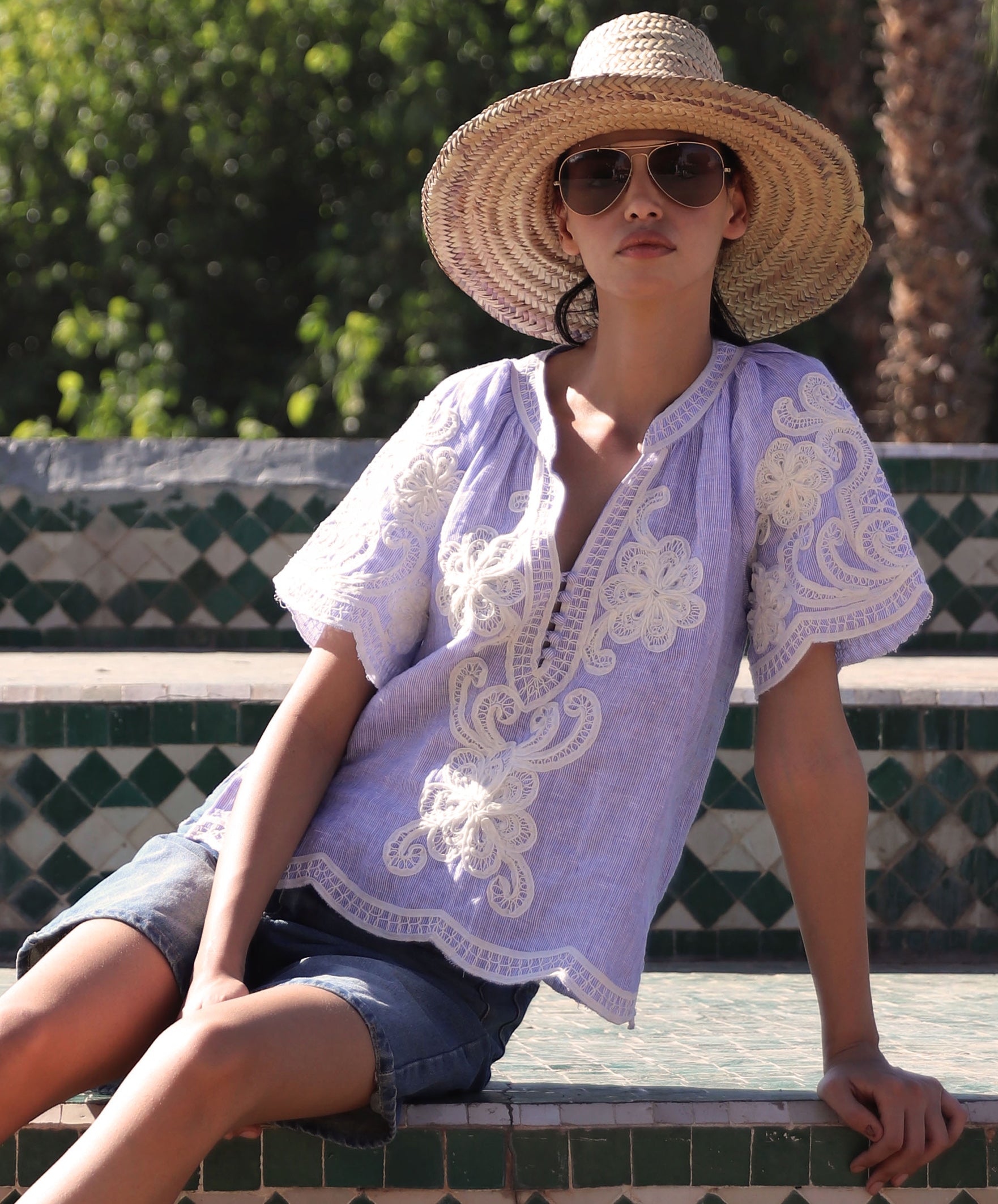 Woman sat on steps wearing a blue striped embroidered Azalea top and straw hat