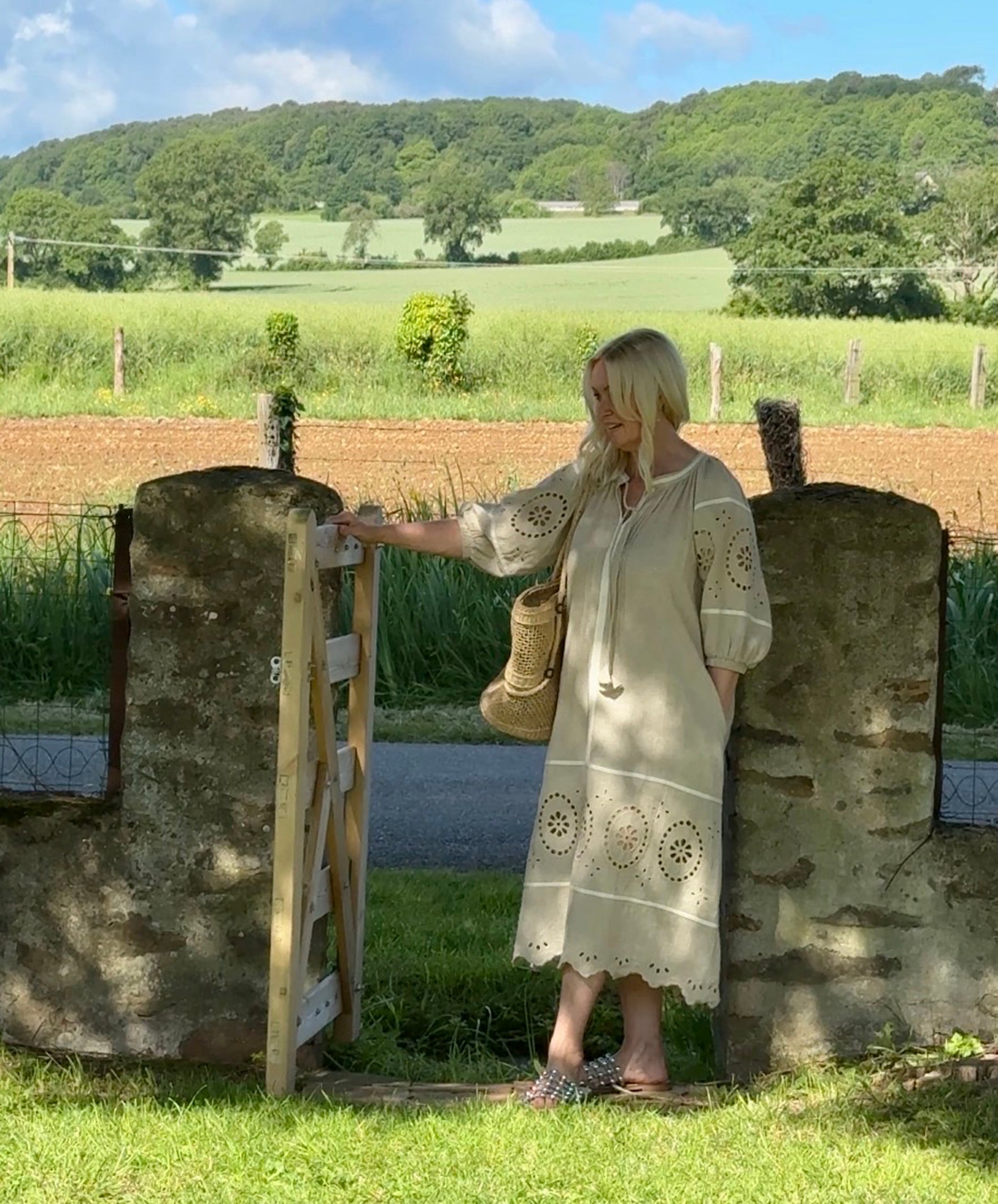 Influencer @shutters_and_stone stood wearing the Magnolia natural embroidered dress standing next to a stone gate with a scenic background of fields and trees.
