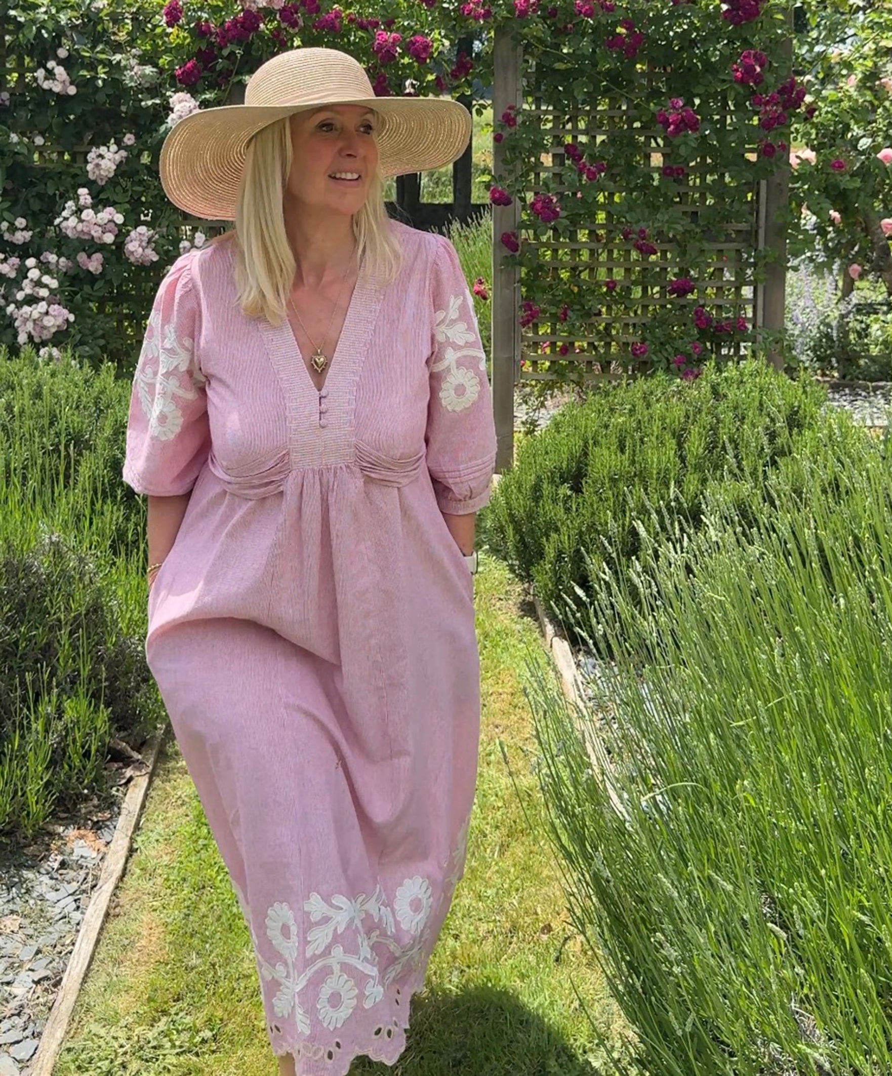 Influencer  @shutters_and_stone wearing the Begonia pink dress and hat standing in a garden with lavender and flowers.